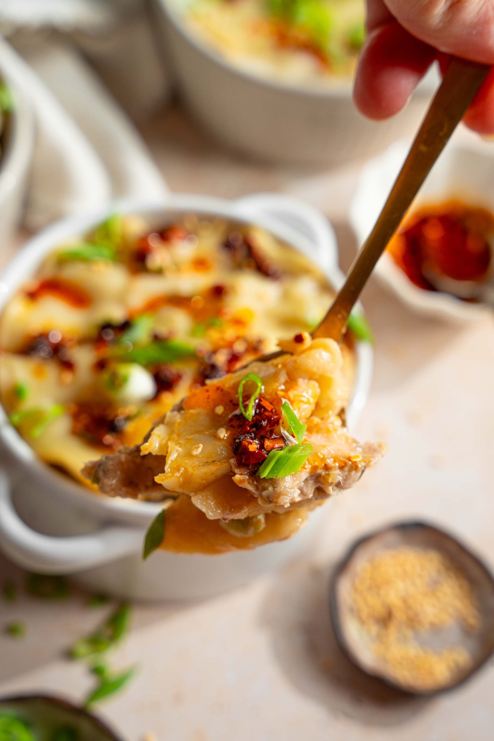 A close up of a fork with a bite of dumpling lasagna garnished with sliced green onion and chili oil. There is a ramekin of lasagna on a tan counter with small plates of garnishes.