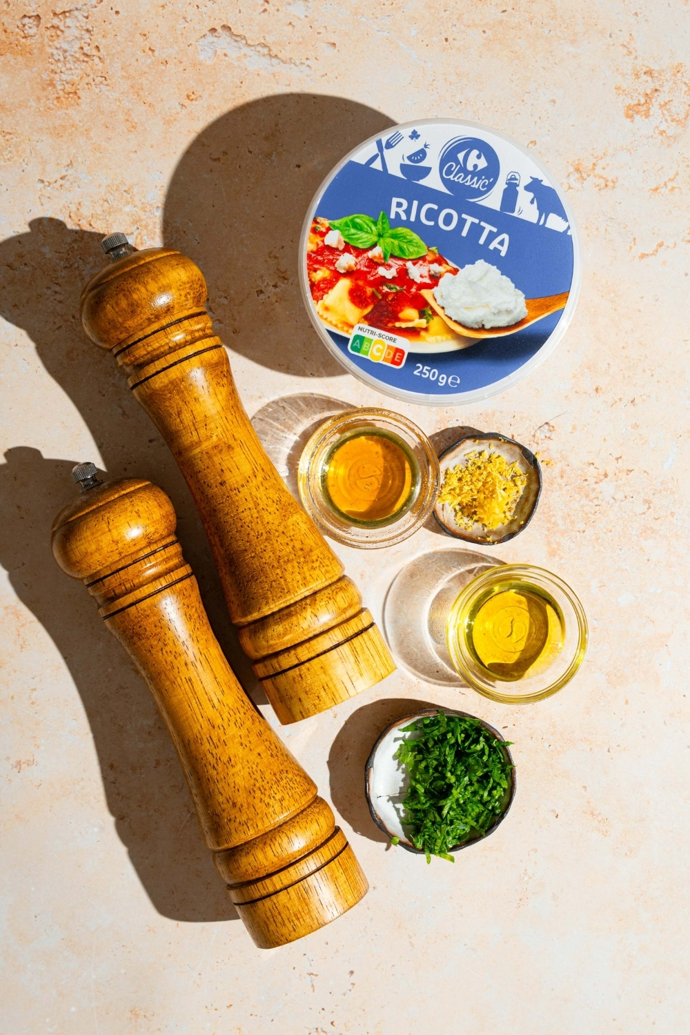 An overhead shot of several bowls in various sizes containing ingredients to make whipped ricotta dip including ricotta cheese, olive oil, honey, herbs, lemon zest, and a salt and pepper mill.