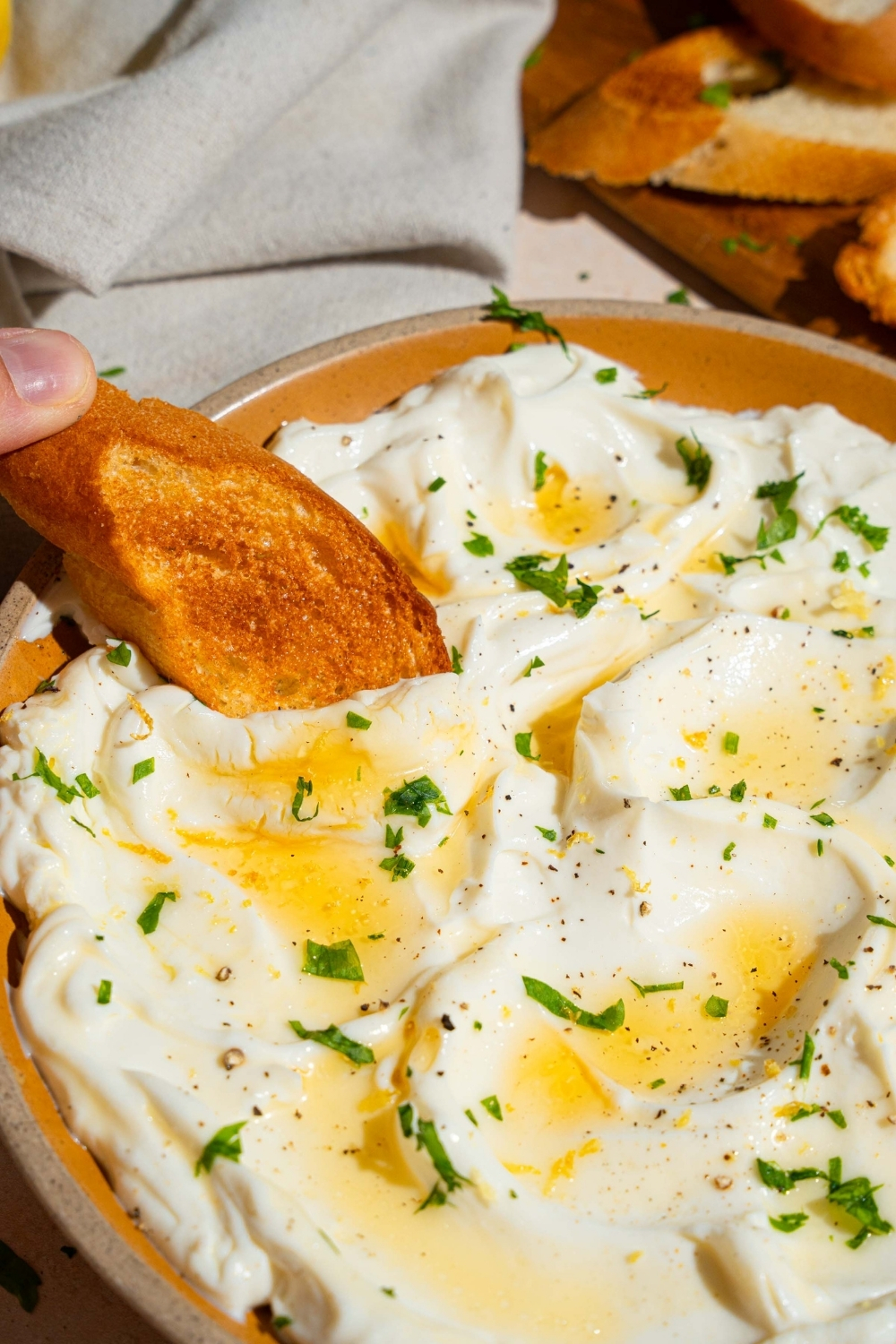 An orange plate with whipped ricotta dip finished with honey and chopped herbs. A slice of toasted baguette is being dipped into the ricotta.