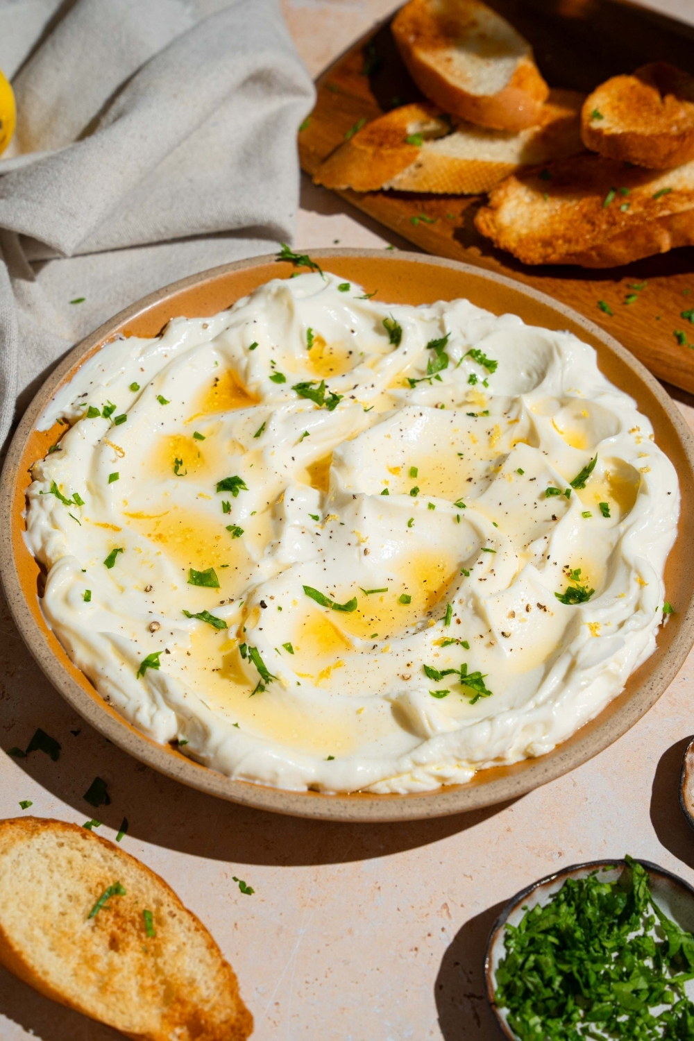 An orange plate with whipped ricotta dip finished with honey and chopped herbs. The plate is on a white counter with a wooden board with toasted baguette slices, and small bowls of garnishes.