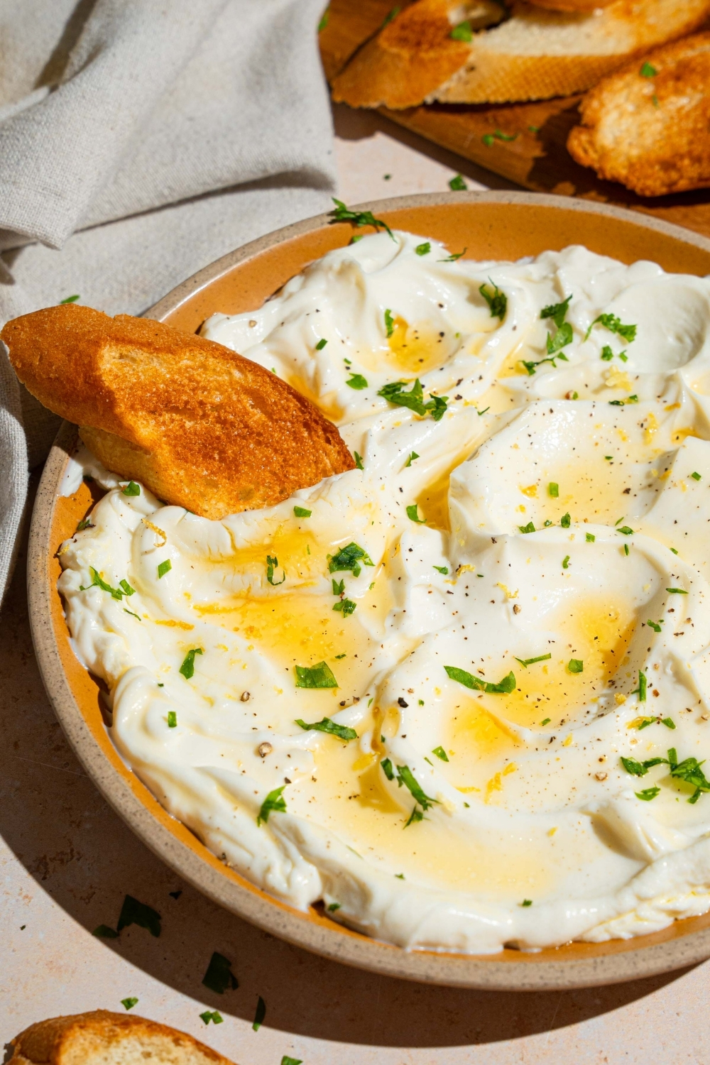 An orange plate with whipped ricotta dip finished with honey and chopped herbs. There is a slice of toasted baguette in the dip. The plate is on a white counter with a wooden board with toasted baguette slices, and small bowls of garnishes.