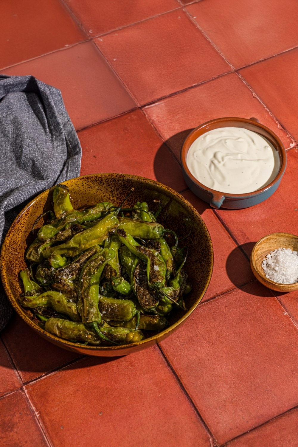 A bowl of blistered shishito peppers with a bowl of creamy lime dip and small bowl of flaky salt on a red tiled counter with a blue cloth napkin.