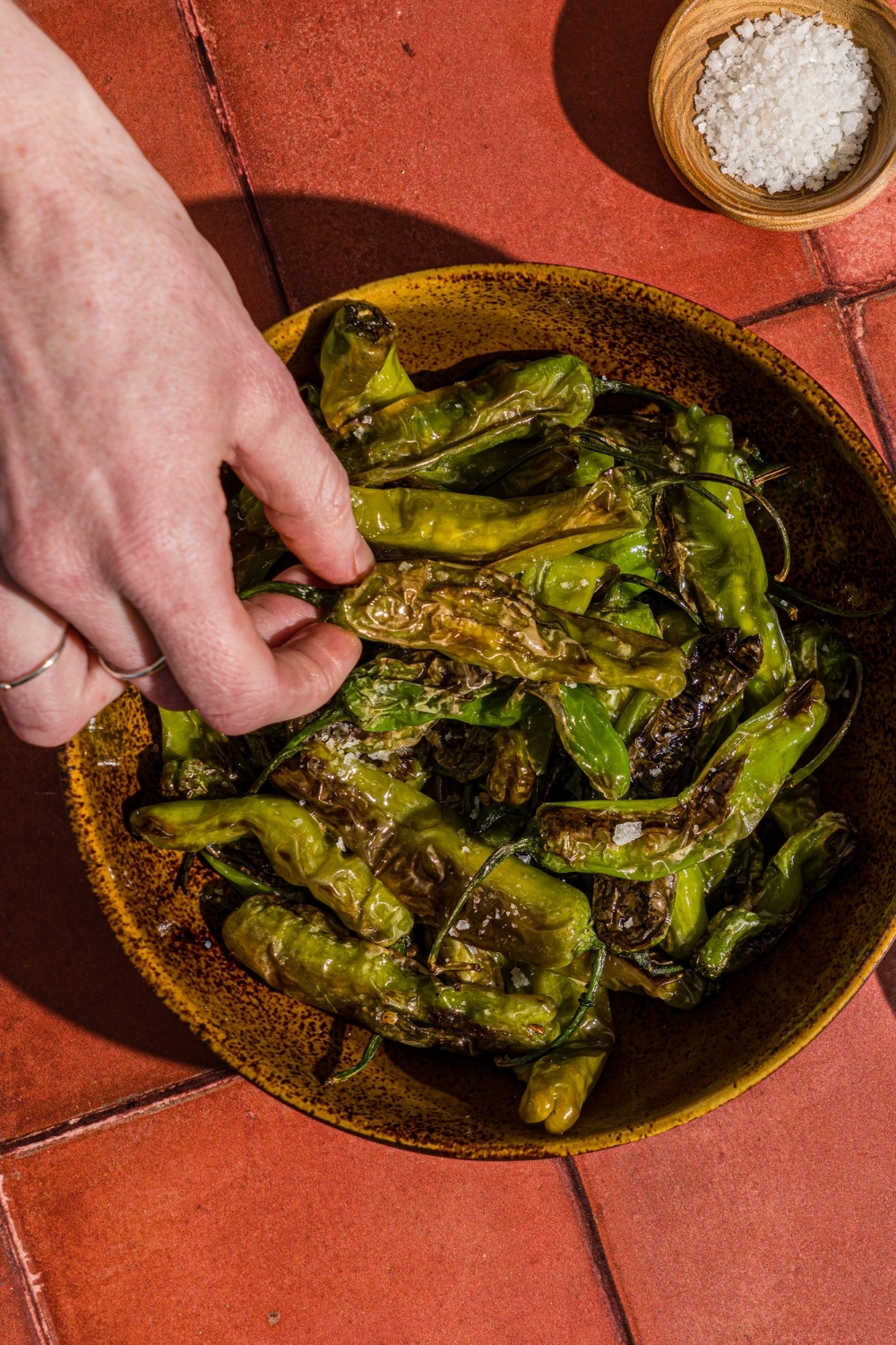 A bowl of blistered shishito peppers with a bowl of flaky salt on a red tiled counter. A hand is taking a pepper from the bowl.