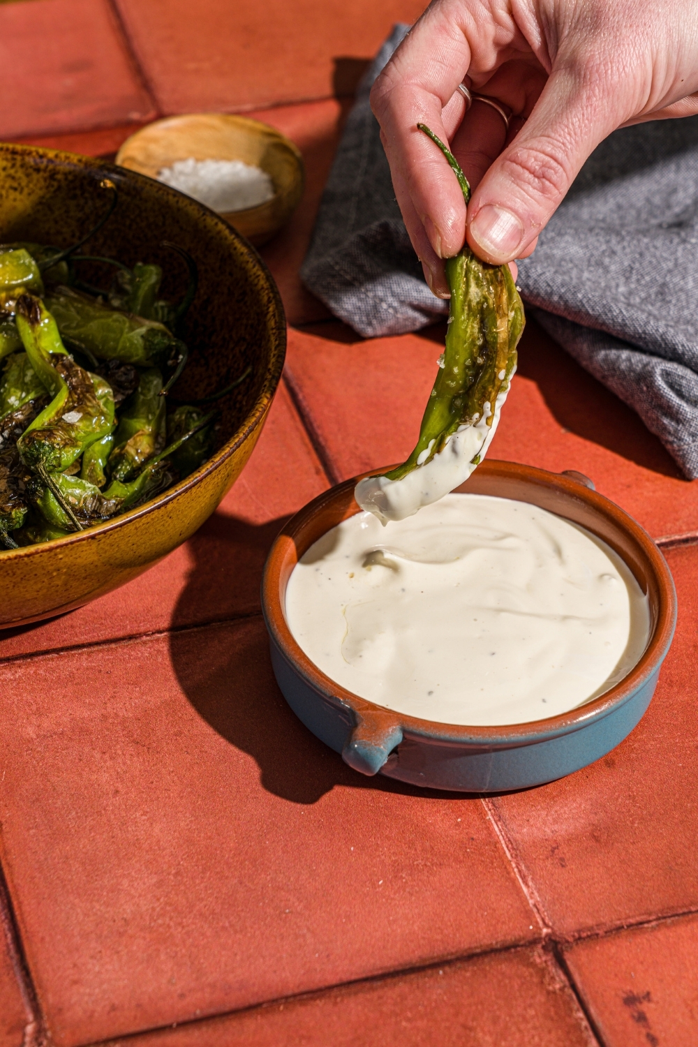 A hand dipping a blistered shishito pepper into a bowl of creamy lime dip. The bowl is on a red tiled counter with a bowl of peppers and blue cloth napkin.
