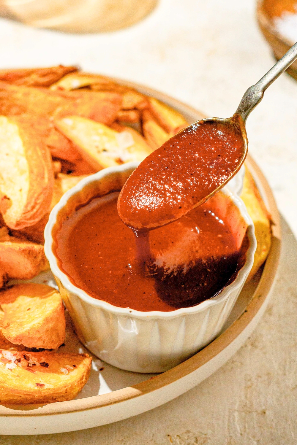 A spoon with Carolina BBQ on it hovering over a bowl of the sauce. The bowl of sauce is on a plate with potato wedges.