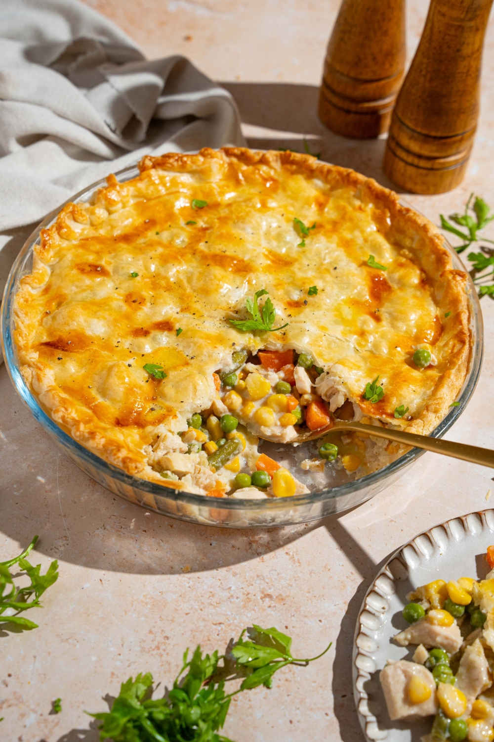 A whole chicken pot pie garnished with fresh parsley with a slice of pie removed. A spoon is in the pie dish. The dish is on a tan counter with a salt and pepper mill and plate with a slice of pot pie.