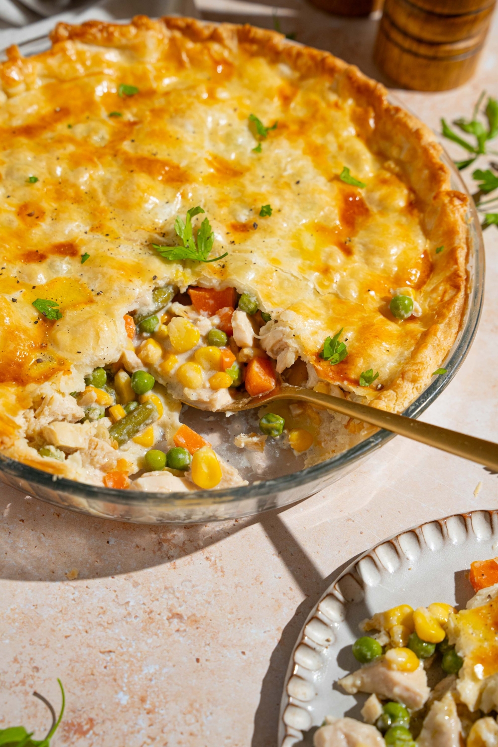A whole chicken pot pie garnished with fresh parsley with a slice of pie removed. A spoon is in the pie dish. The dish is on a tan counter with a plate with a slice of pot pie.