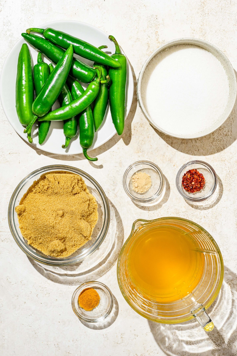 A plate of jalapenos, a bowl of sugar, a bowl of brown sugar, a small bowl of turmeric, a small bowl of garlic powder, a small bowl of aleppo pepper, and a bowl of apple cider vinegar.