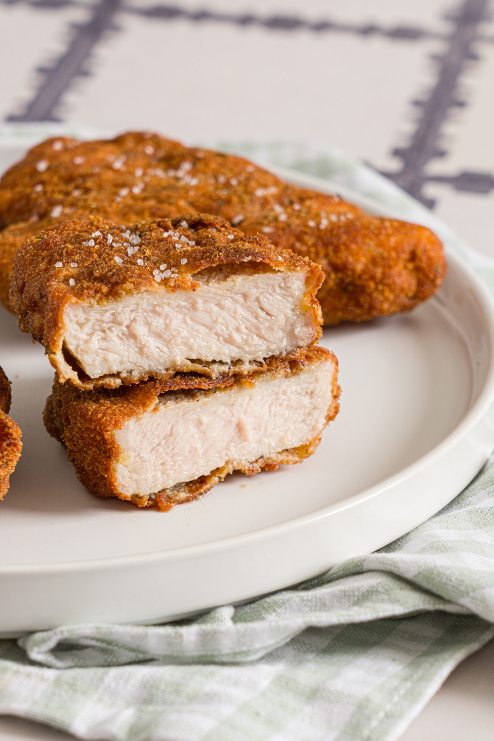 A white plate with crispy breaded pork cutlets. One cutlet is sliced in half and stacked on top of each other. The plate is on a tiled counter with a green cloth napkin.