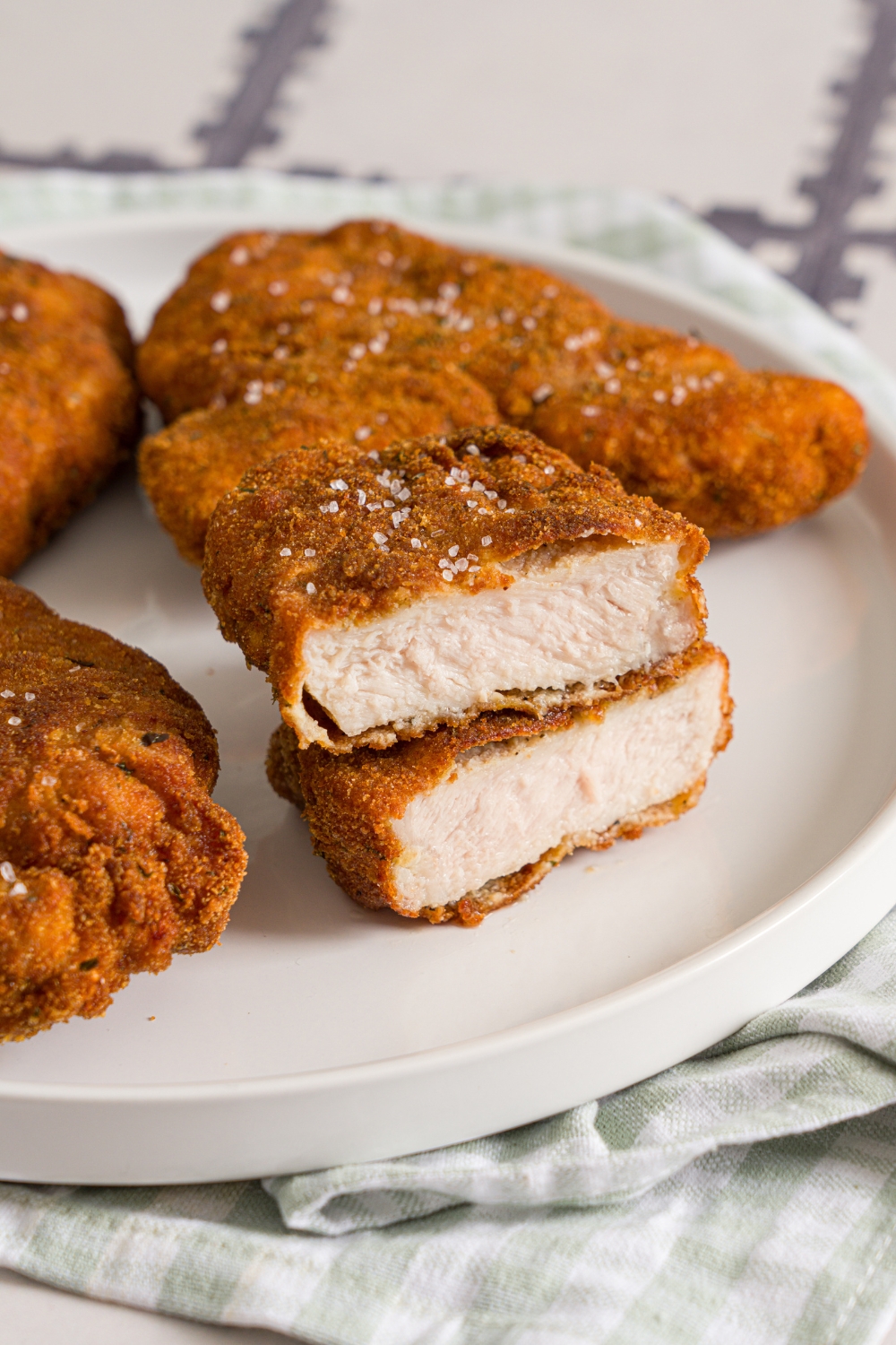 A white plate with crispy breaded pork cutlets. One cutlet is sliced in half and stacked on top of each other. The plate is on a tiled counter with a green cloth napkin.
