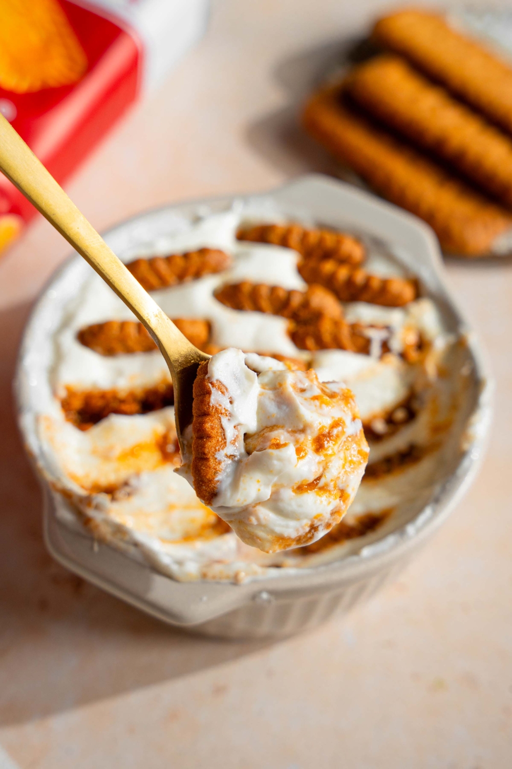 A spoon that has Biscoff cookies and yogurt on it being held above a ramekin that is filled with the Biscoff yogurt cheesecake mixture.