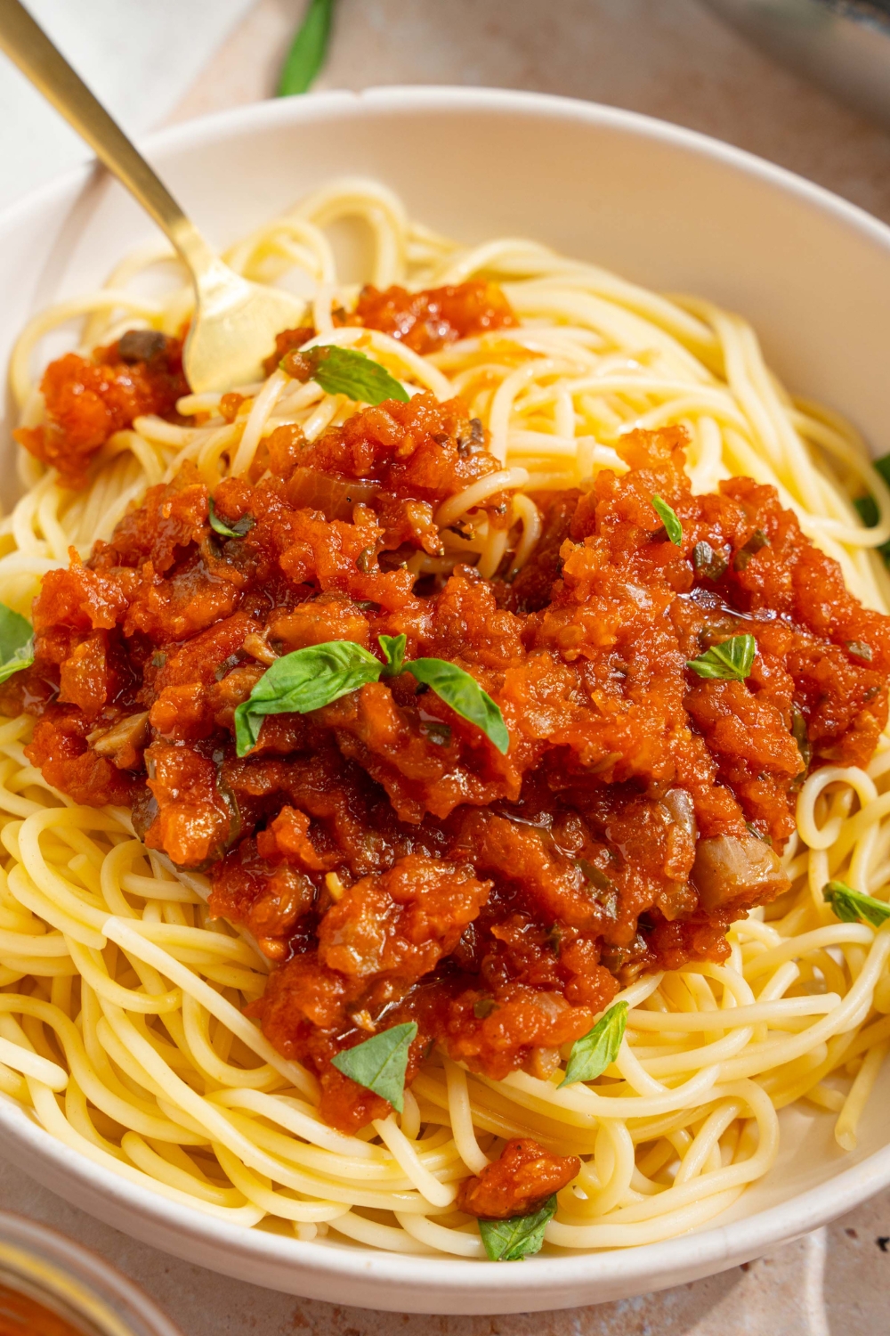A bowl of spaghetti with homemade spaghetti sauce with fresh tomatoes. The bowl is garnished with fresh basil. There is a fork in the bowl. The bowl is on a tan counter with a white cloth napkin.
