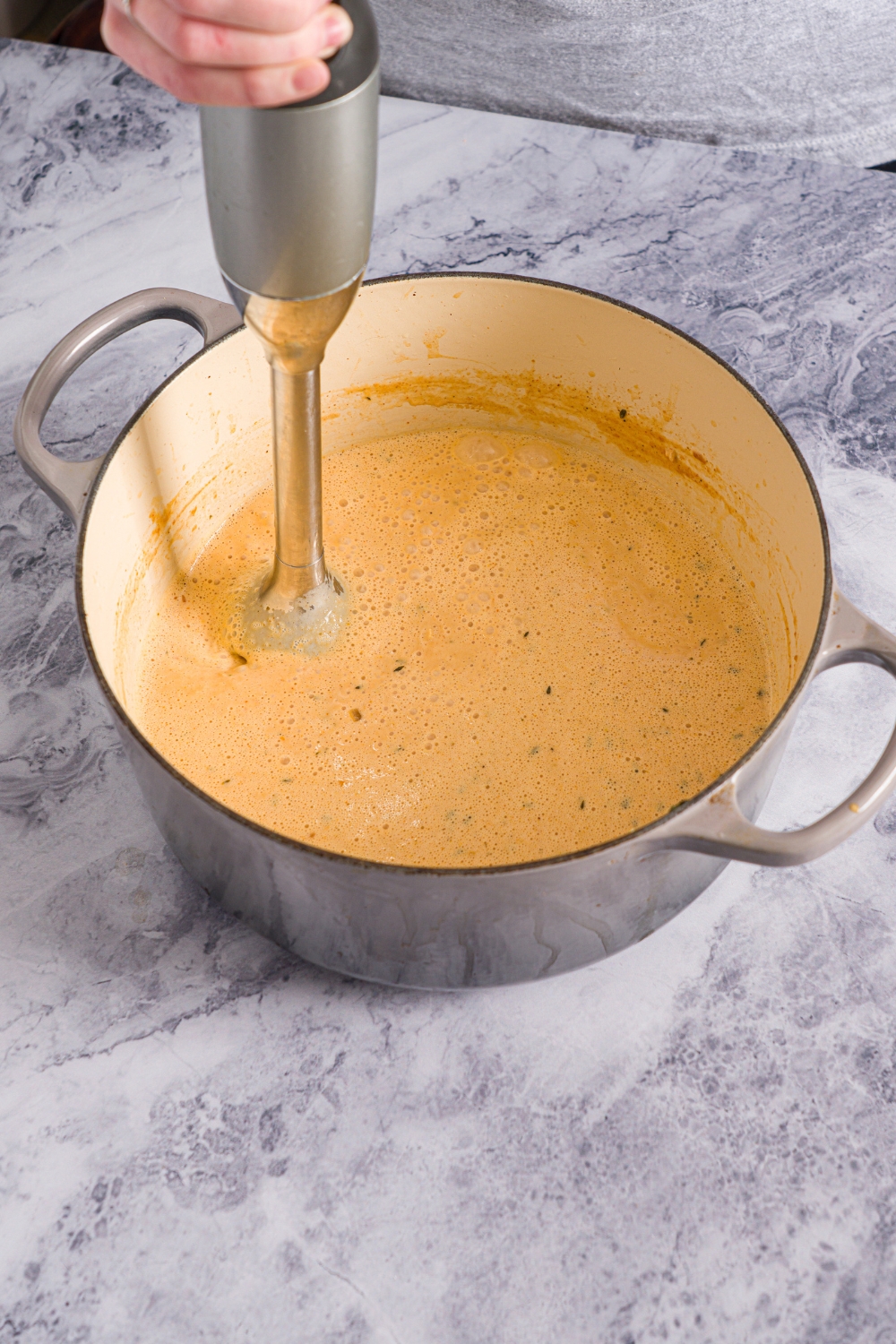 A dutch oven with an immersion blender mixing a shrimp bisque base. The pot is on a marble counter.