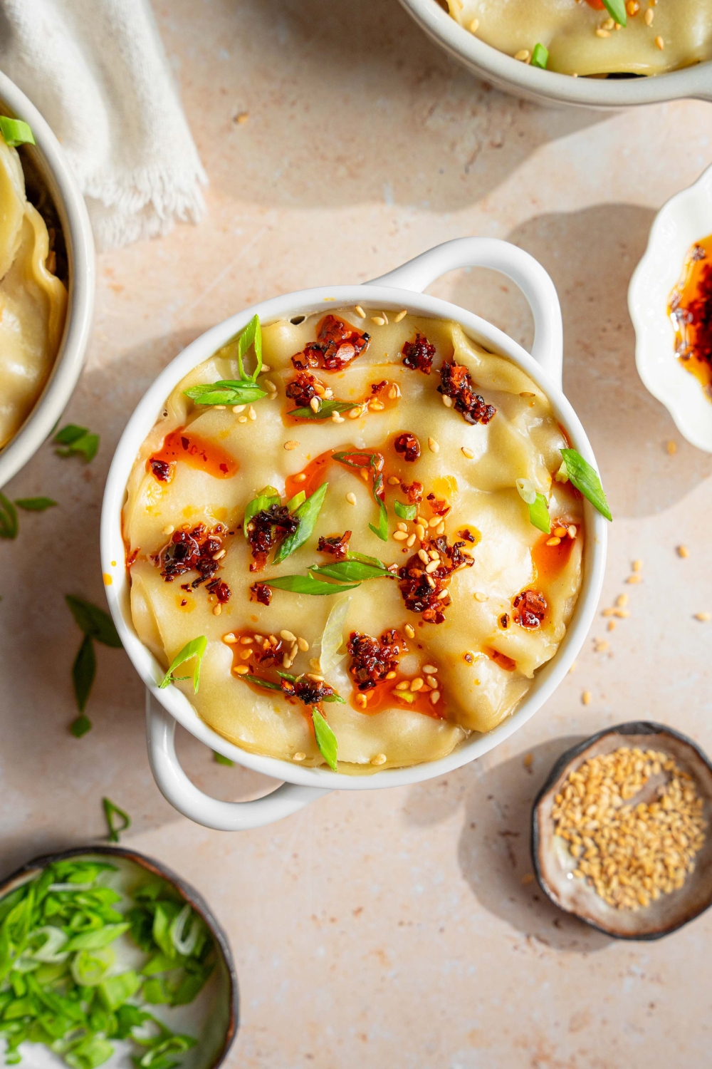 A ramekin with dumpling lasagna topped with chili oil, sliced green onions, and sesame seeds. The ramekin is on a tan counter with additional ramekins of dumpling lasagna, a white cloth napkin, and a small bowls of garnishes.