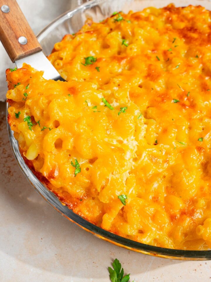 A round glass baking dish with baked 3 cheese mac and cheese garnished with fresh parsley. The dish is on a tan counter with a spatula in the mac and cheese.