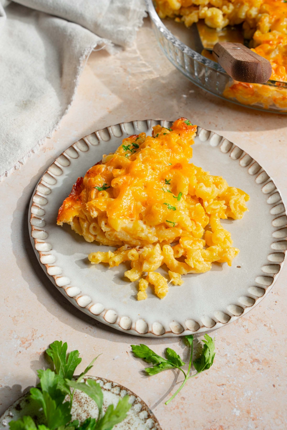 A plate with 3 cheese mac and cheese garnished with fresh parsley. The plate is on a tan counter with a baking dish with mac and cheese, white cloth napkin, and small plate of garnishes.