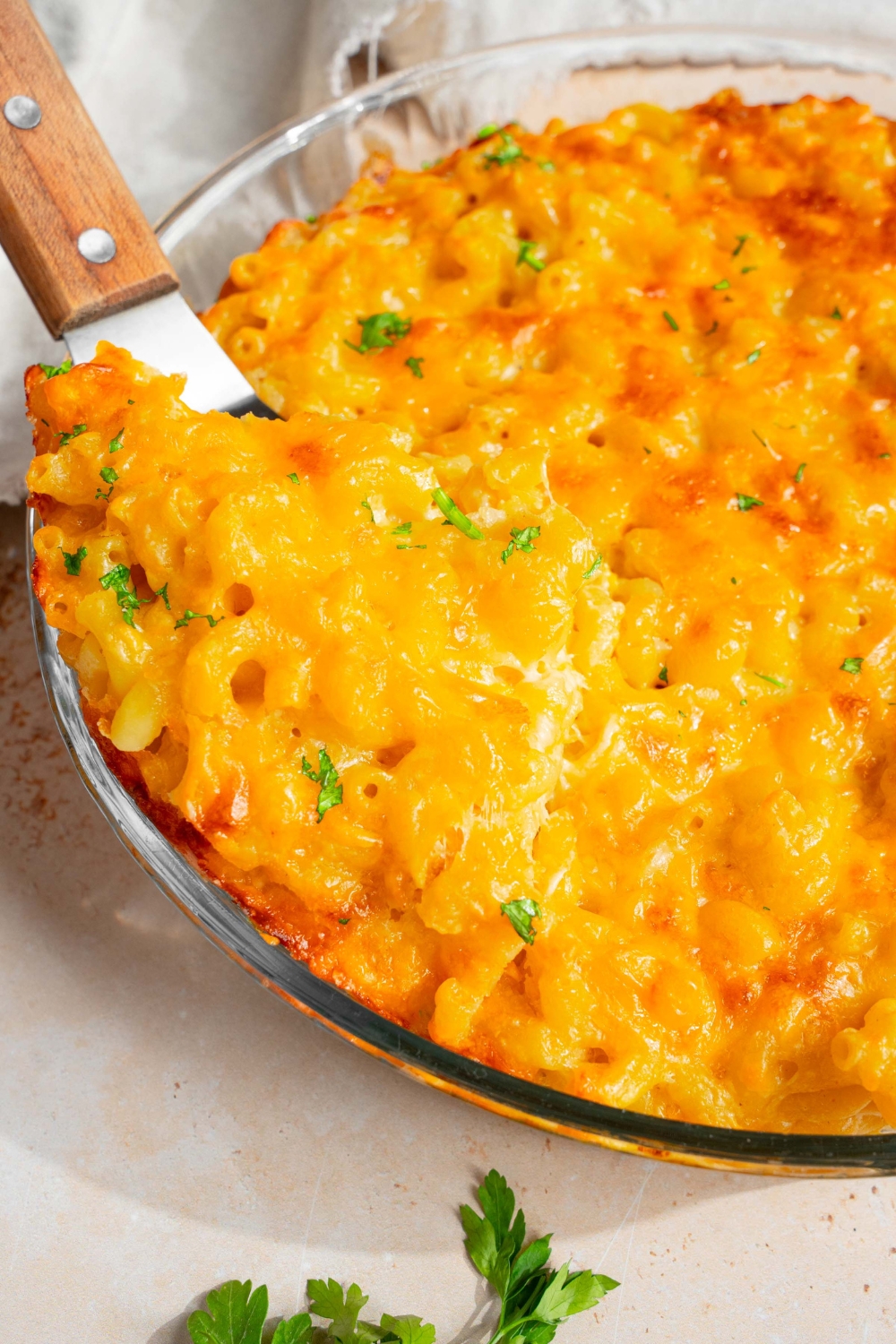 A round glass baking dish with baked 3 cheese mac and cheese garnished with fresh parsley. The dish is on a tan counter with a spatula in the mac and cheese.