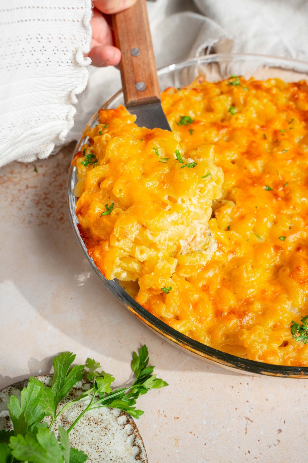 A round glass baking dish with baked 3 cheese mac and cheese garnished with fresh parsley. The dish is on a tan counter with a spatula in the mac and cheese.