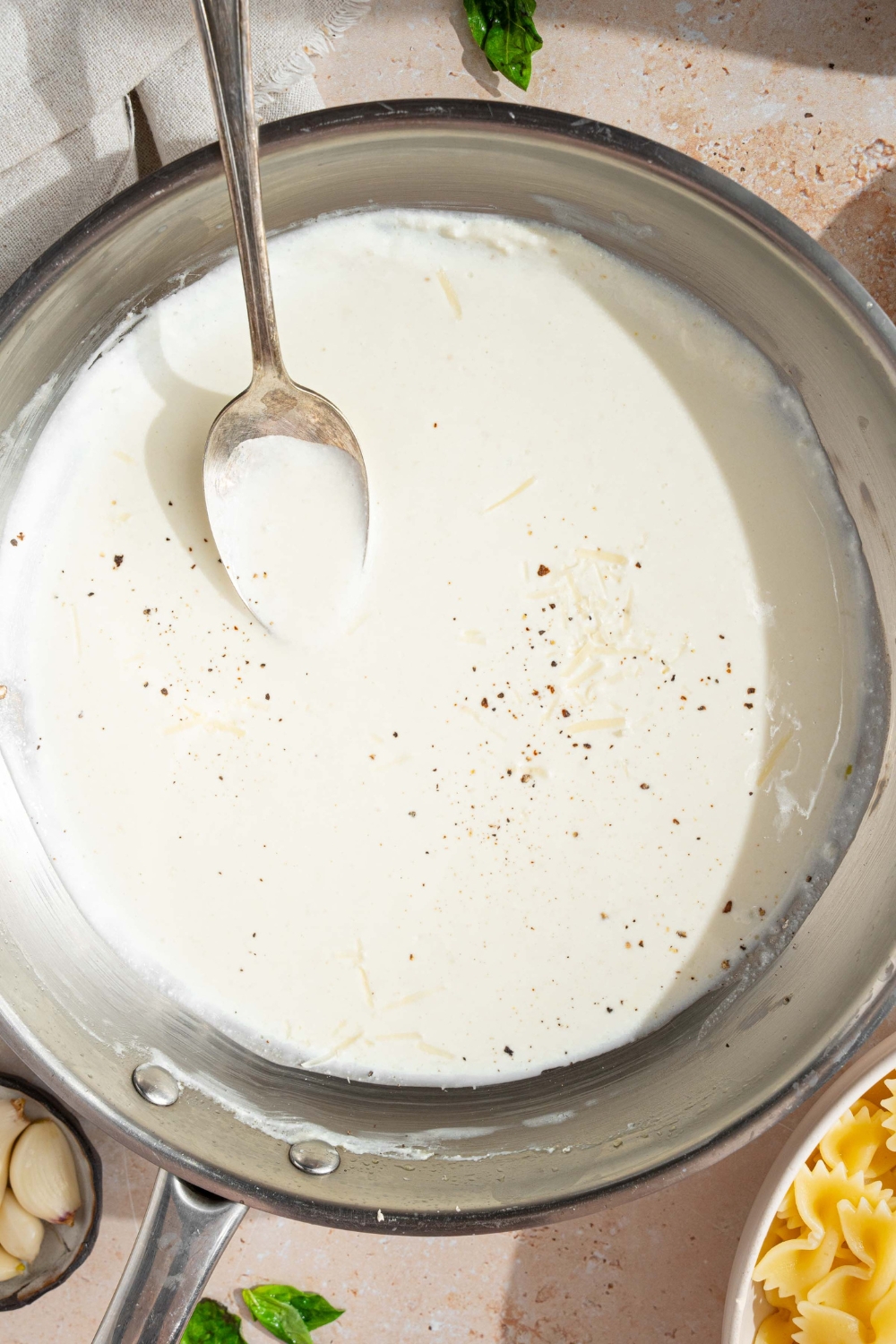 A skillet with alfredo sauce with cream cheese finished with cracked black pepper. A spoon is stirring the sauce. The skillet is on a tan counter with a bowl of bowtie pasta.