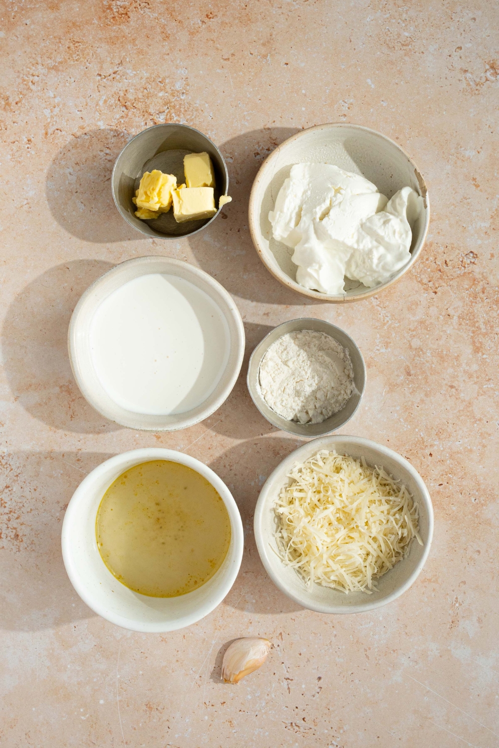 An overhead shot of several bowls in various sizes containing ingredients to make alfredo sauce including cream cheese, butter, chicken stock, heavy cream, and shredded parmesan cheese.
