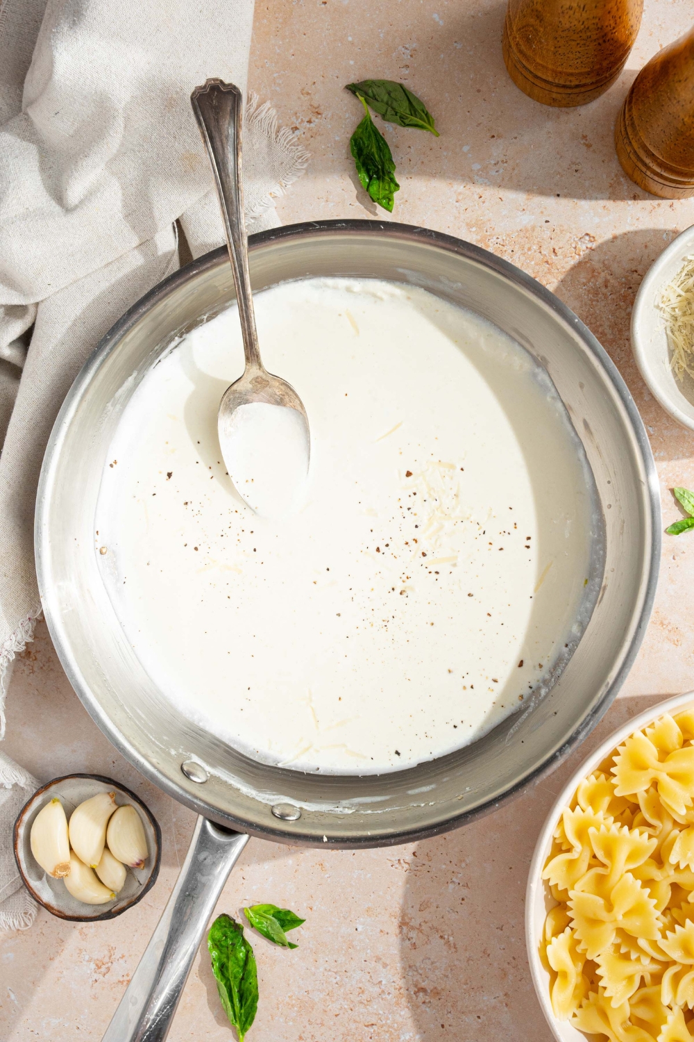 A skillet with alfredo sauce with cream cheese finished with cracked black pepper. A spoon is in the sauce. The skillet is on a tan counter with a white cloth napkin, small bowl of garlic, and bowl of bowtie pasta.