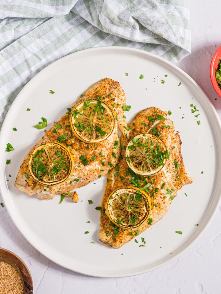 A white plate with two baked red snapper fillets with a garlic topping and topped with sliced lemon and fresh parsley. The plate is on a white counter with a green checkered napkin and small bowl of garnishes.