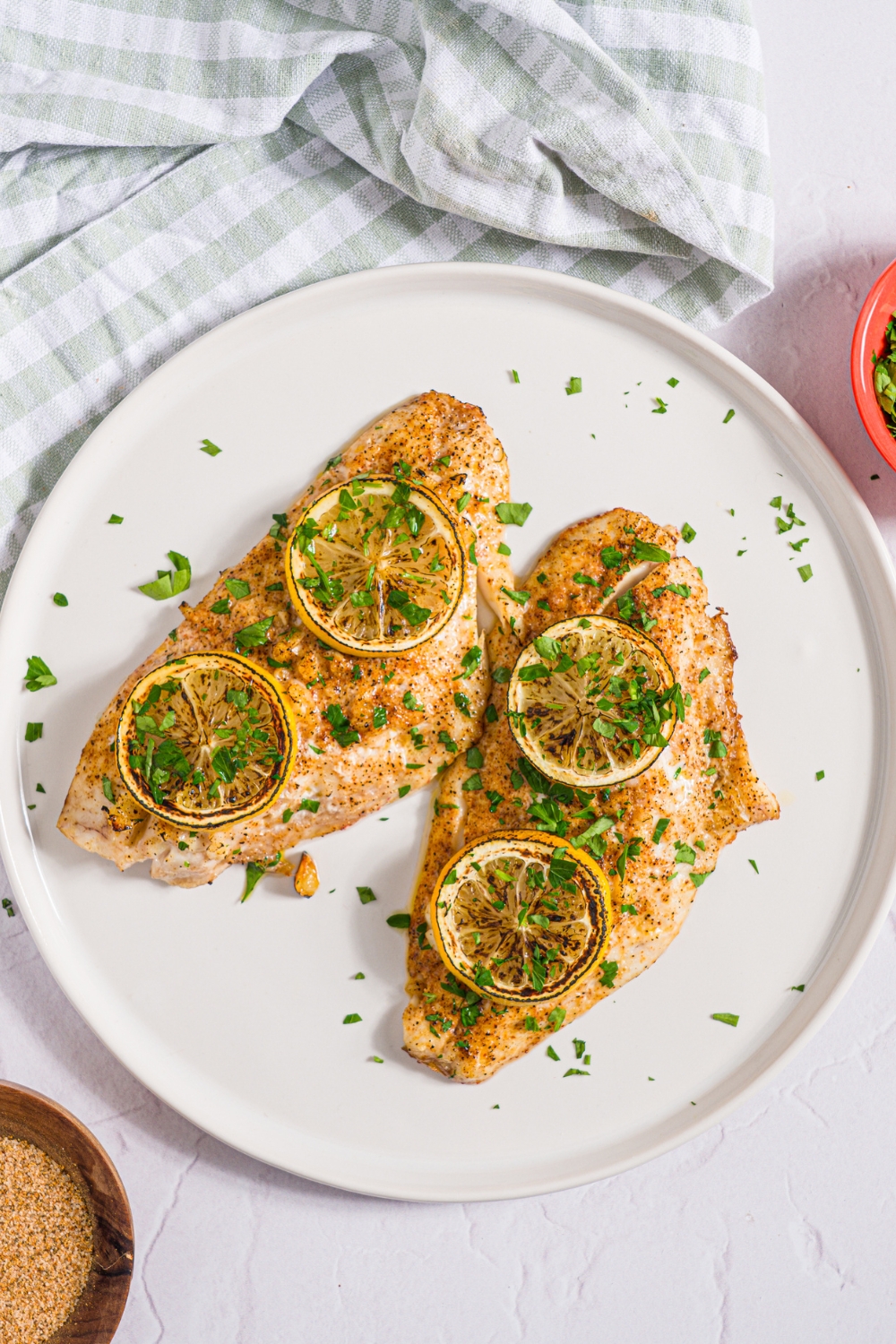 A white plate with two baked red snapper fillets with a garlic topping and topped with sliced lemon and fresh parsley. The plate is on a white counter with a green checkered napkin and small bowl of garnishes.