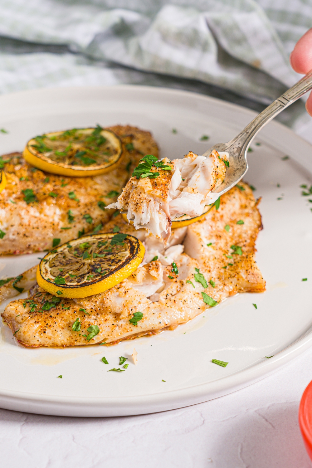 A white plate with two baked red snapper fillets with a garlic topping and topped with sliced lemon and fresh parsley. A fork is taking a bite of snapper. The plate is on a white counter with a green checkered napkin.