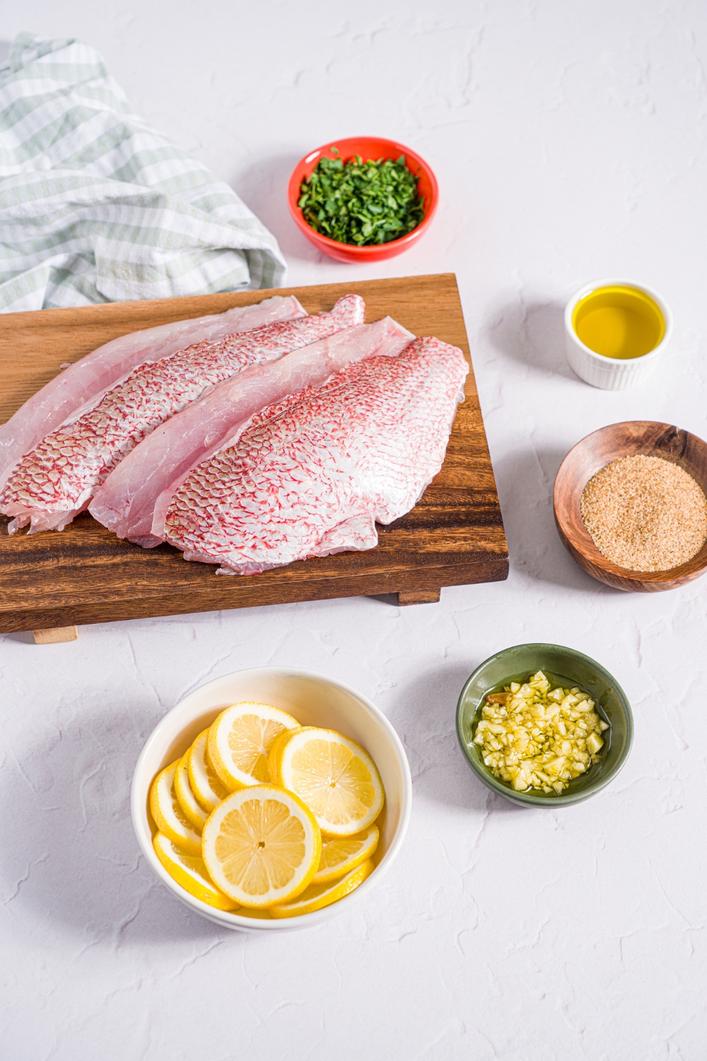 A white counter with ingredients to make baked red snapper including red snapper fillets on a wooden board, and small bowls of sliced lemon, garlic, parsley, olive oil, and creole seasoning.