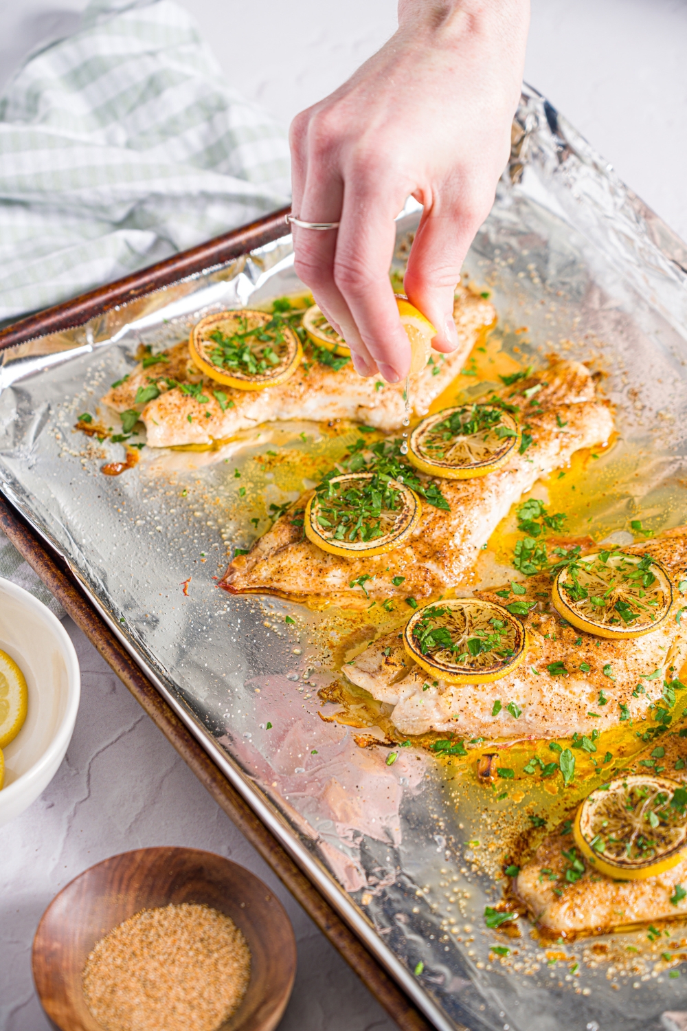 A baking sheet lined with foil with baked red snapper with a garlic topping and sliced lemon. A hand is squeezing lemon over the fish. The sheet is on a white counter with a green checkered napkin and small bowls of garnishes.