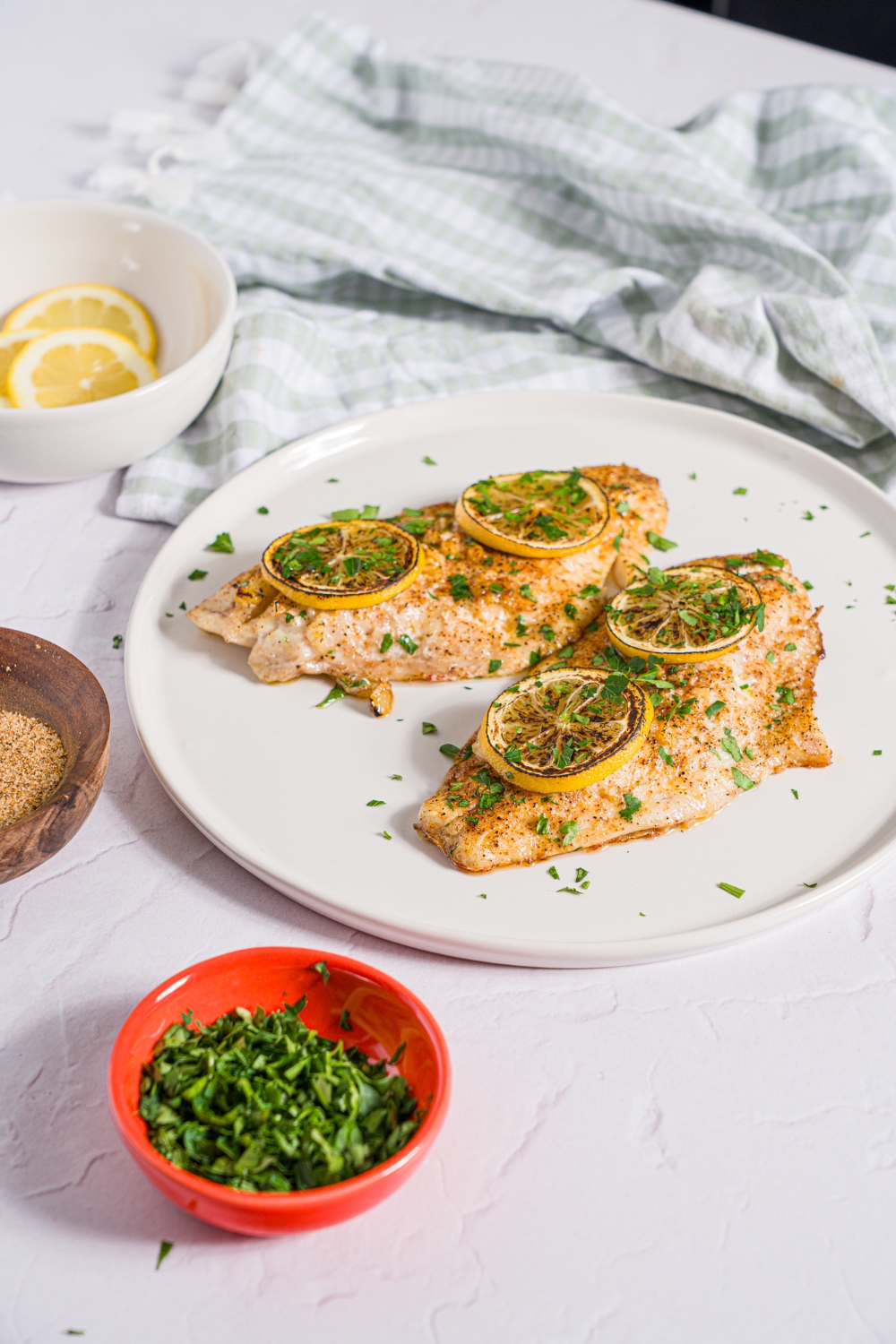 A white plate with two baked red snapper fillets with a garlic topping and topped with sliced lemon and fresh parsley. The plate is on a white counter with a green checkered napkin and small bowl of garnishes.