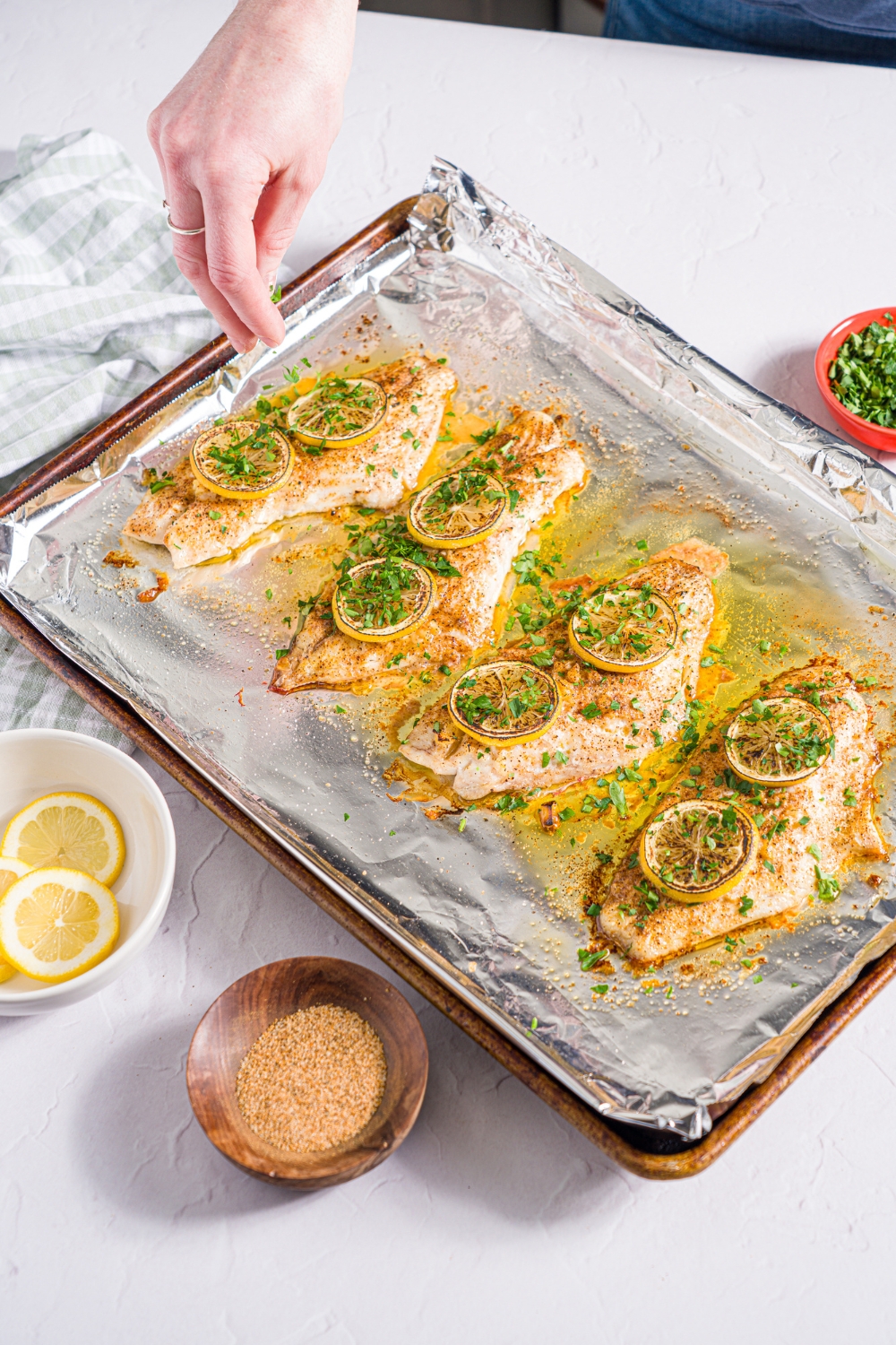 A baking sheet lined with foil with baked red snapper with a garlic topping and sliced lemon. A hand is sprinkling fresh parsley over the fish. The sheet is on a white counter with a green checkered napkin and small bowls of garnishes.