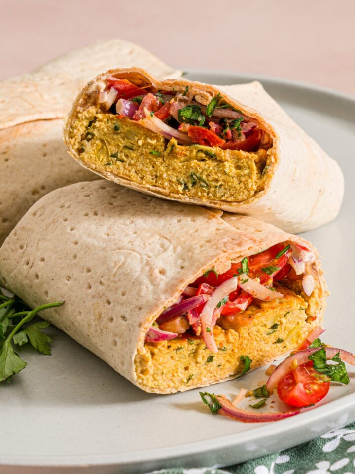 A white plate with two baking sheet chicken kefta wraps. One of the wraps is sliced in half and stacked. The plate is garnished with fresh parsley. The plate is on a tan counter with a green floral napkin.