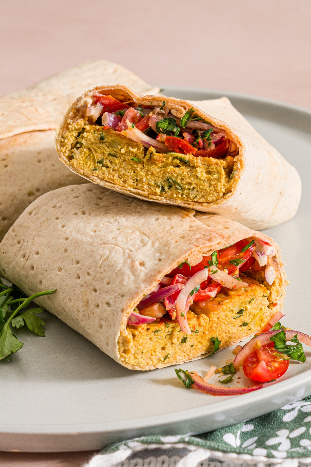 A white plate with two baking sheet chicken kefta wraps. One of the wraps is sliced in half and stacked. The plate is garnished with fresh parsley. The plate is on a tan counter with a green floral napkin.