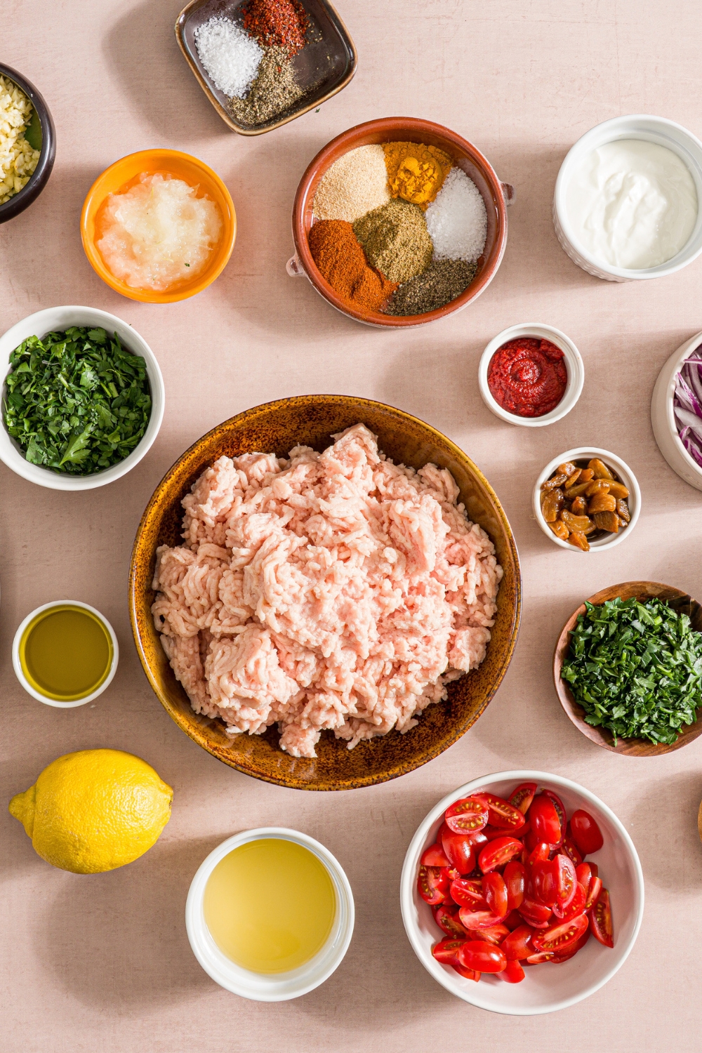 An overhead shot of several bowls in various sizes containing ingredients to make baking sheet chicken kefta wraps including ground chicken, parsley, olive oil, tomatoes, lemon, tomato paste, garlic, greek yogurt, onion, and seasonings.