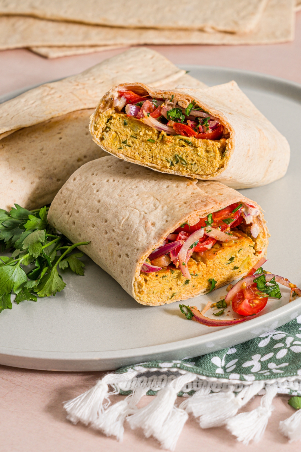 A white plate with two baking sheet chicken kefta wraps. One of the wraps is sliced in half and stacked. The plate is garnished with fresh parsley. The plate is on a tan counter with a green floral napkin.