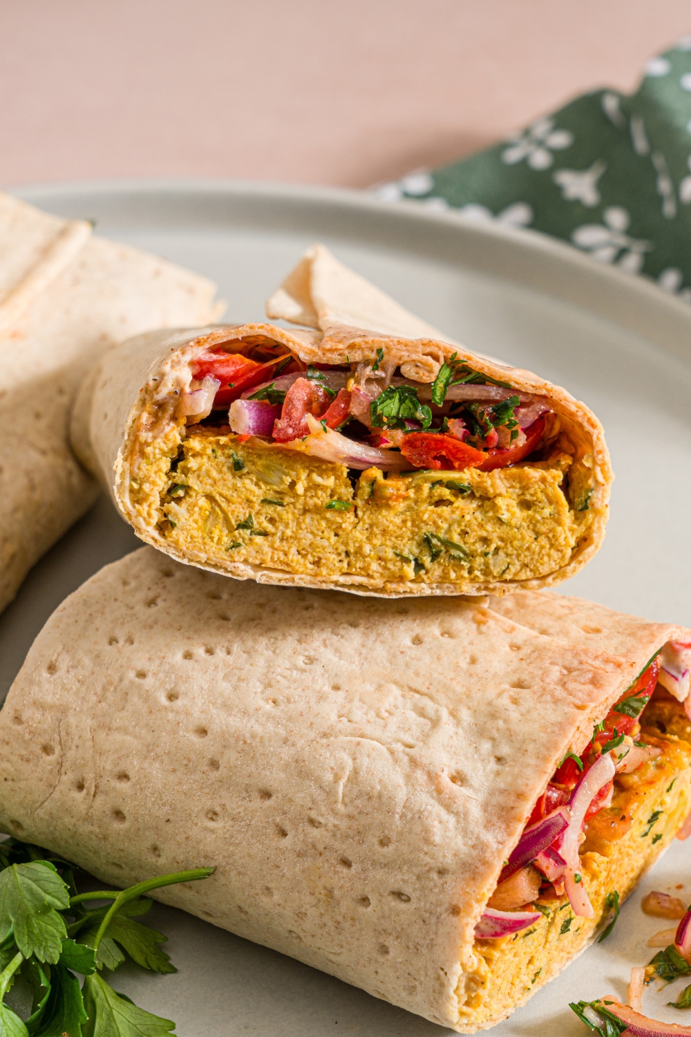 A white plate with two baking sheet chicken kefta wraps. One of the wraps is sliced in half and stacked. The plate is garnished with fresh parsley. The plate is on a tan counter with a green floral napkin.