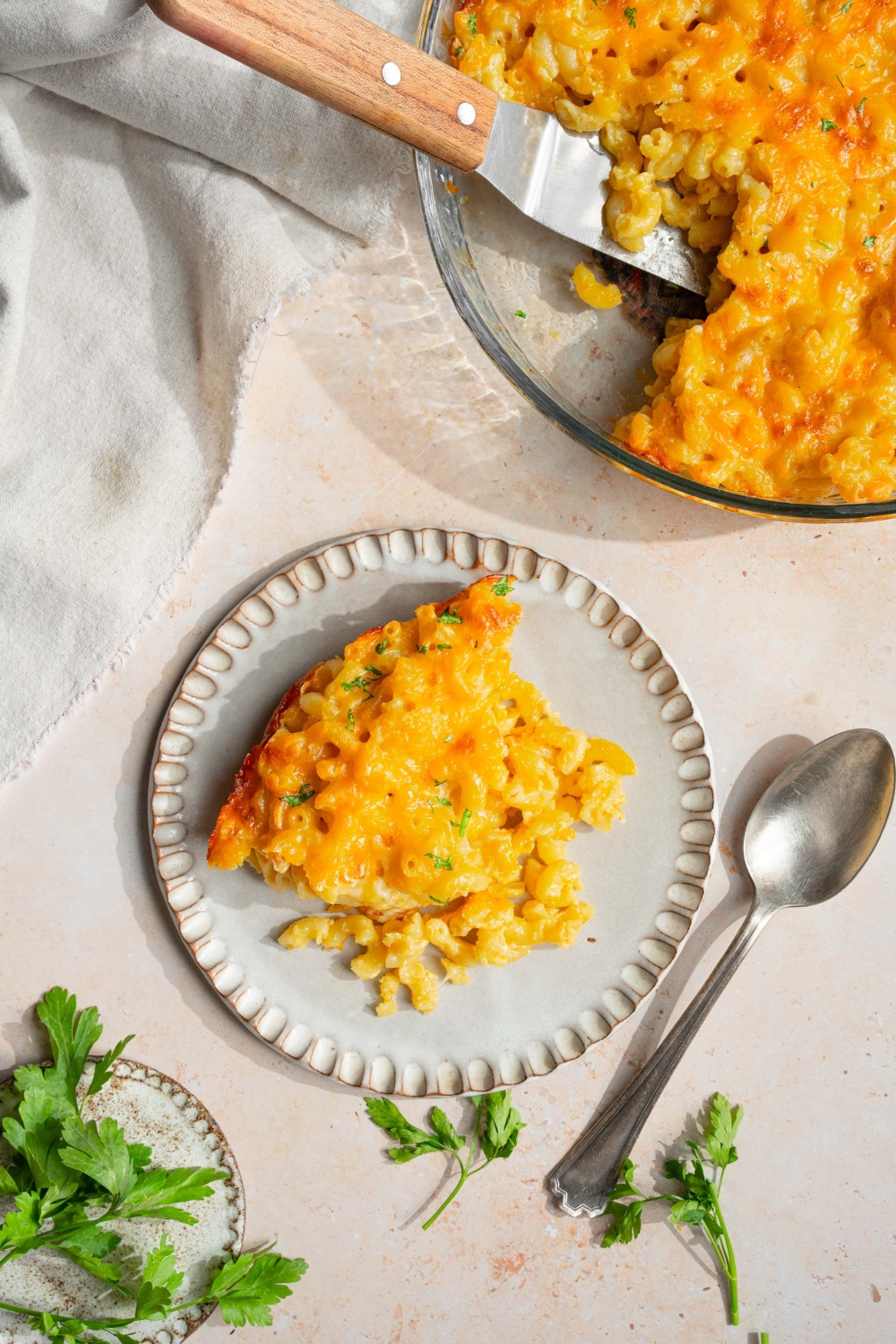 A plate with 3 cheese mac and cheese garnished with fresh parsley. The plate is on a tan counter with a baking dish with mac and cheese, a spoon, a white cloth napkin, and small plate of garnishes.