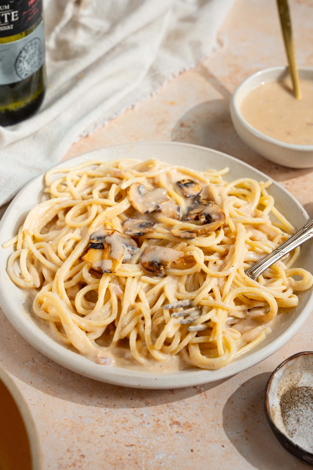 A white plate with spaghetti tossed in sherry cream sauce topped with mushrooms. A fork is in the spaghetti. The plate is on a tan counter with a bowl of cream sauce and bottle of sherry.