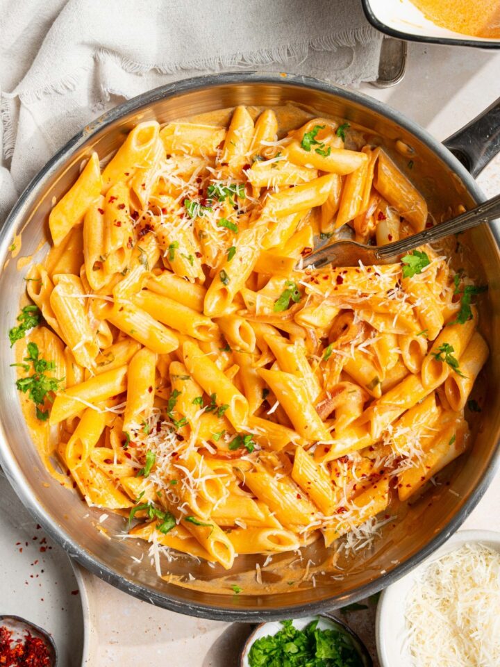 A skillet with pink sauce pasta garnished with shredded parmesan cheese, fresh parsley, and crushed red pepper. There is a fork in the pasta. The skillet is on a tan counter with a small bowl of cheese, small plates of garnishes, and white cloth napkin.