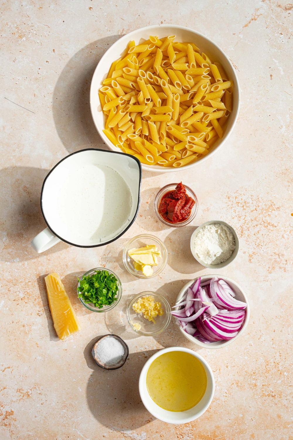 An overhead shot of several bowls in various sizes containing ingredients to make blush sauce pasta including penne pasta, half and half, white wine, sliced onion, tomato paste, butter, garlic, salt, parmesan cheese, and fresh parsley.