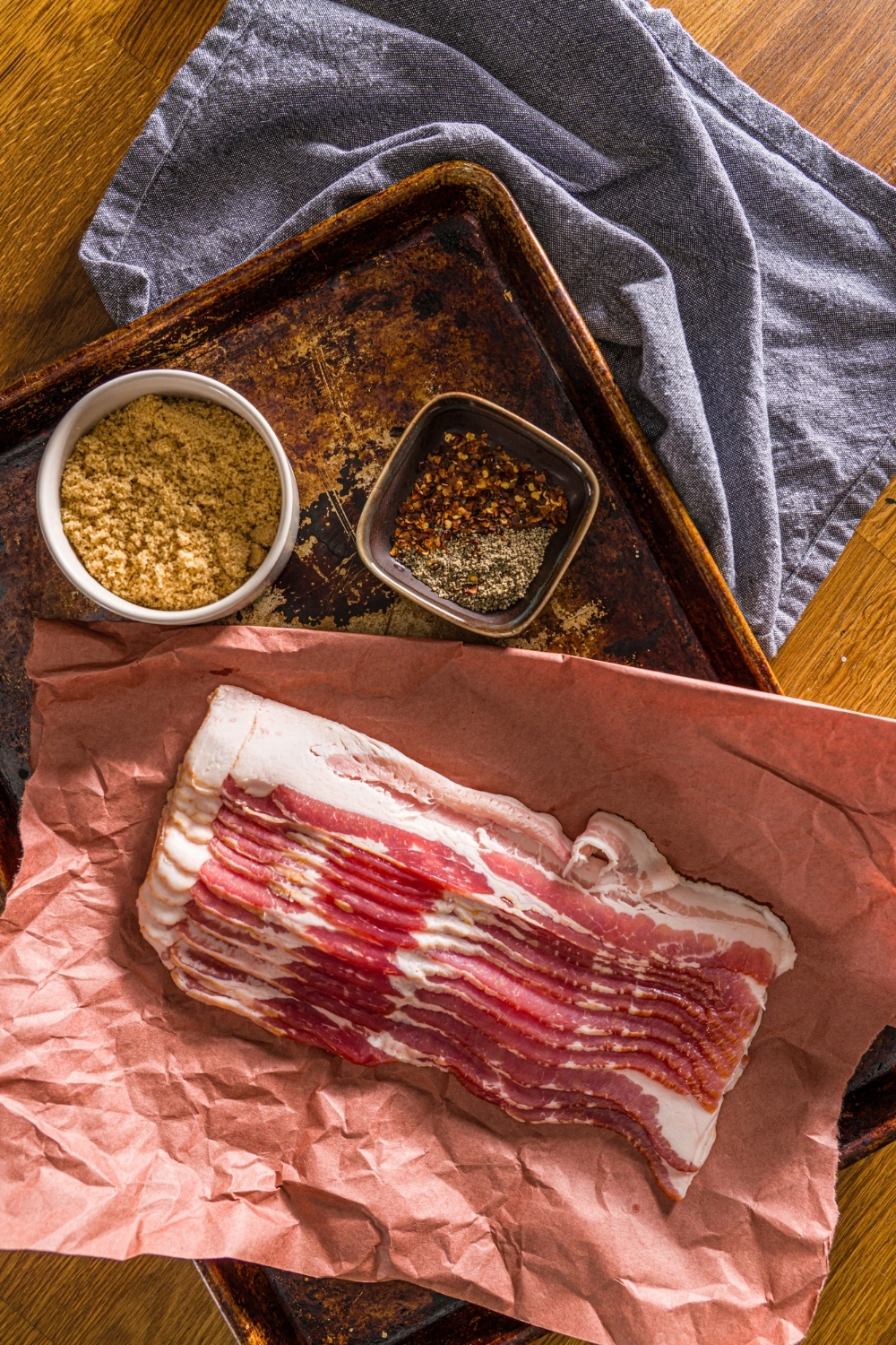 An overhead shot of ingredients to make brown sugar bacon on a baking sheet including bacon, brown sugar, and black pepper. The sheet is on a wooden counter with a blue napkin.