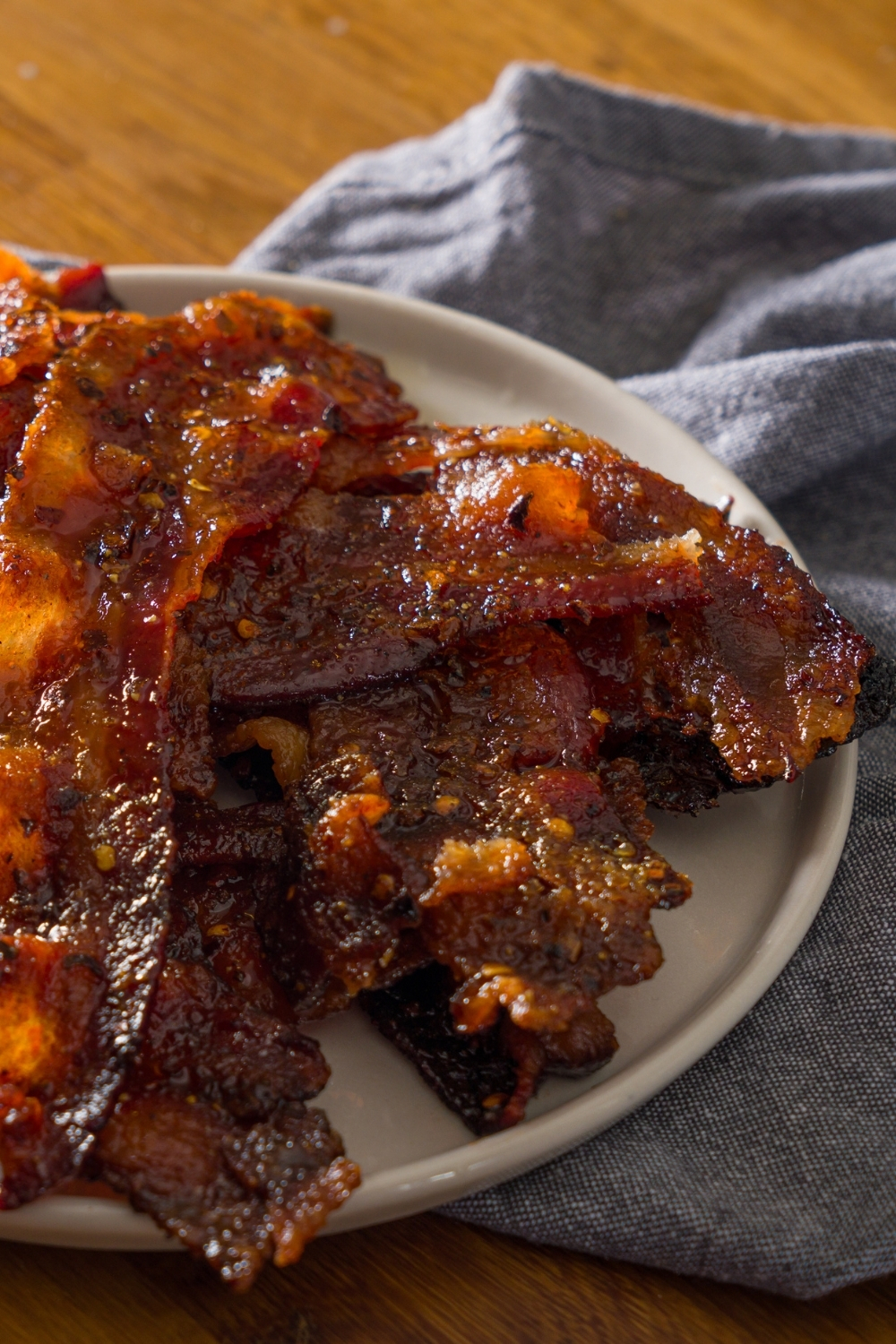 A white plate with brown sugar bacon on a wooden counter with a blue cloth napkin.