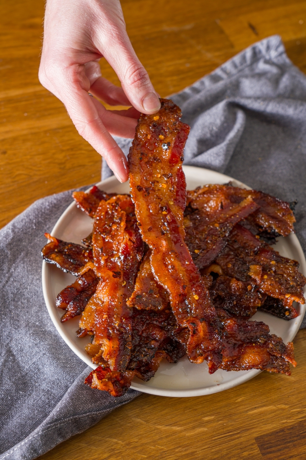 A white plate with brown sugar bacon on a wooden counter with a blue cloth napkin. A hand is taking a strip of bacon.
