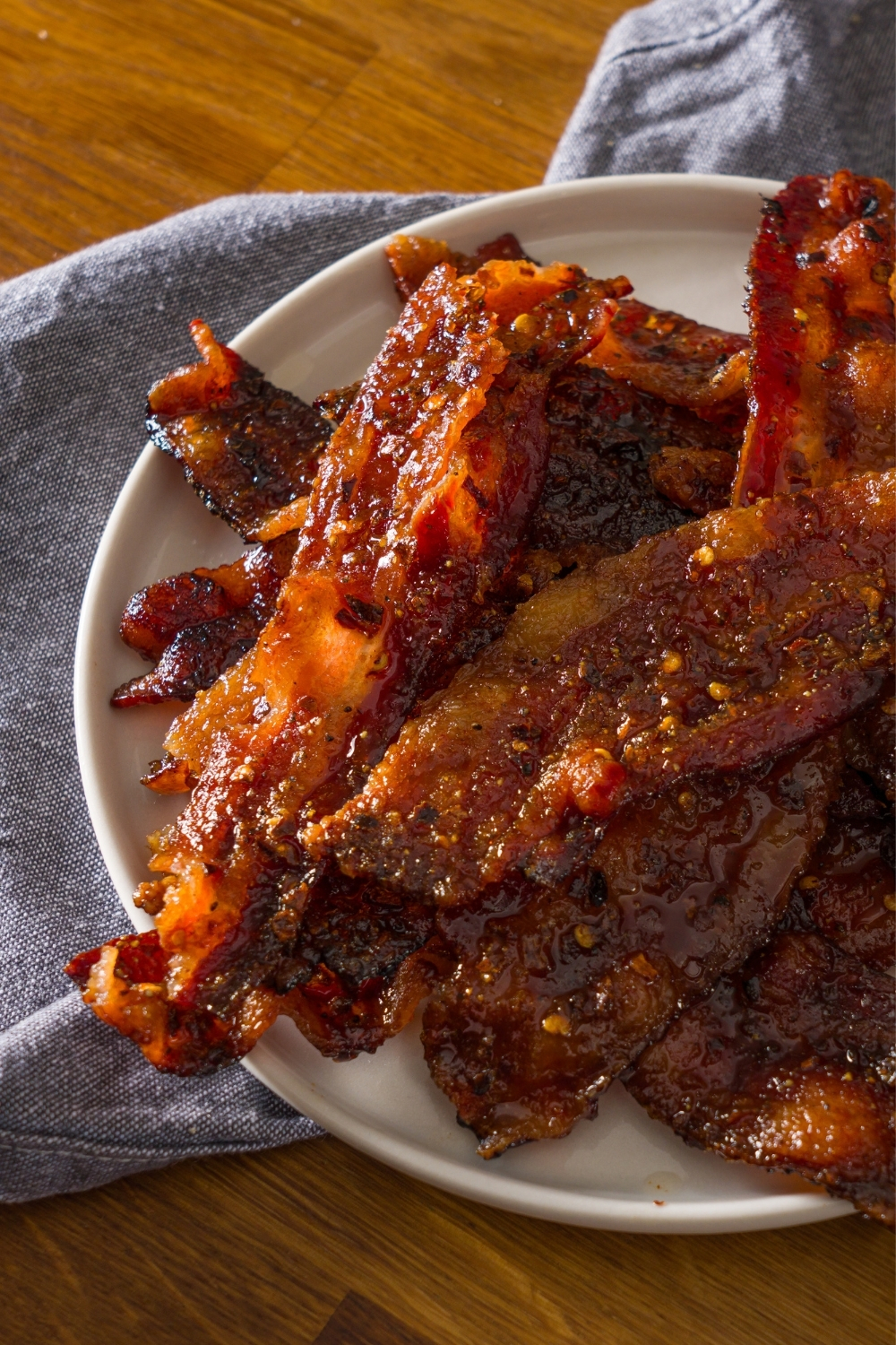 A white plate with brown sugar bacon on a wooden counter with a blue cloth napkin.