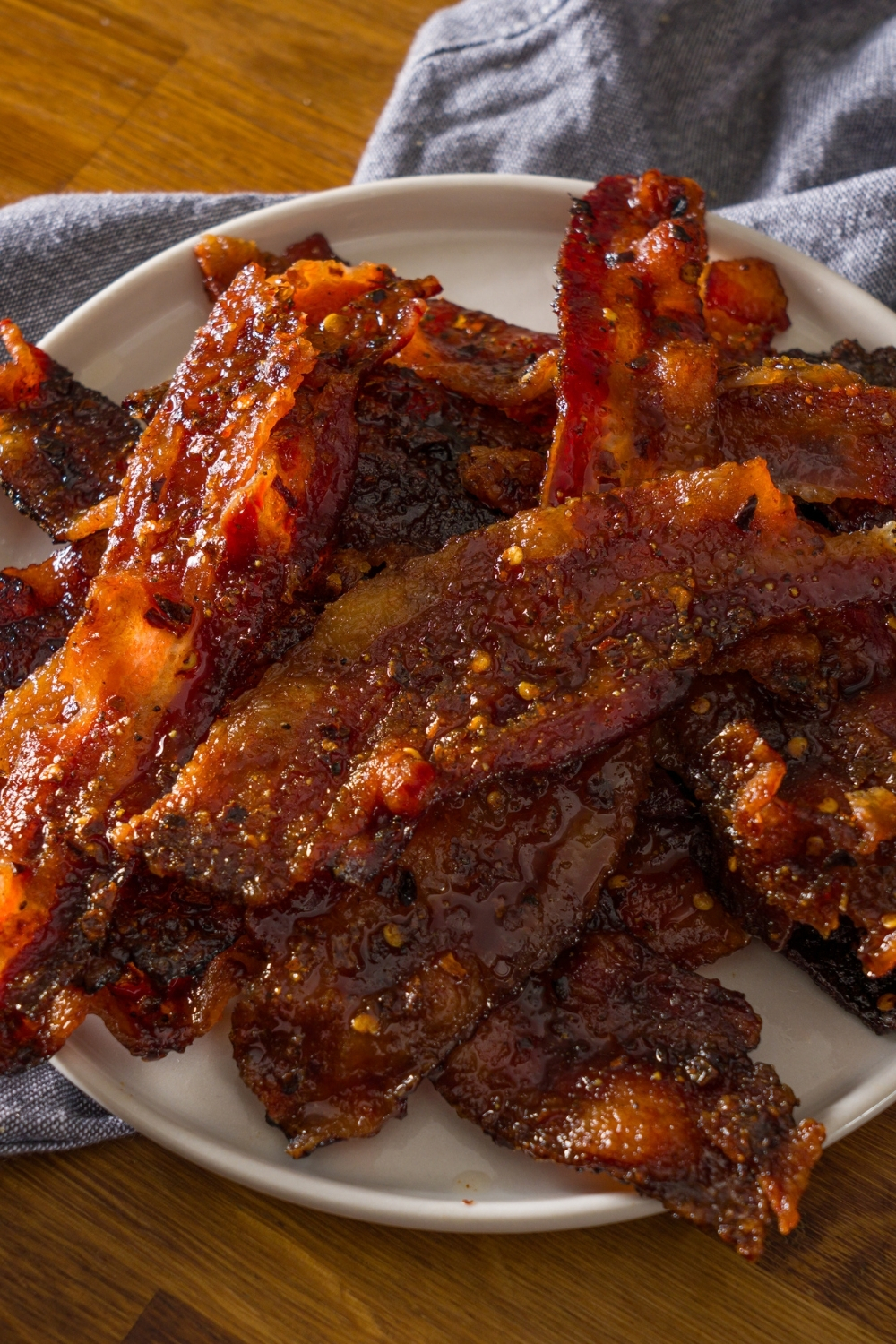 A white plate with brown sugar bacon on a wooden counter with a blue cloth napkin.