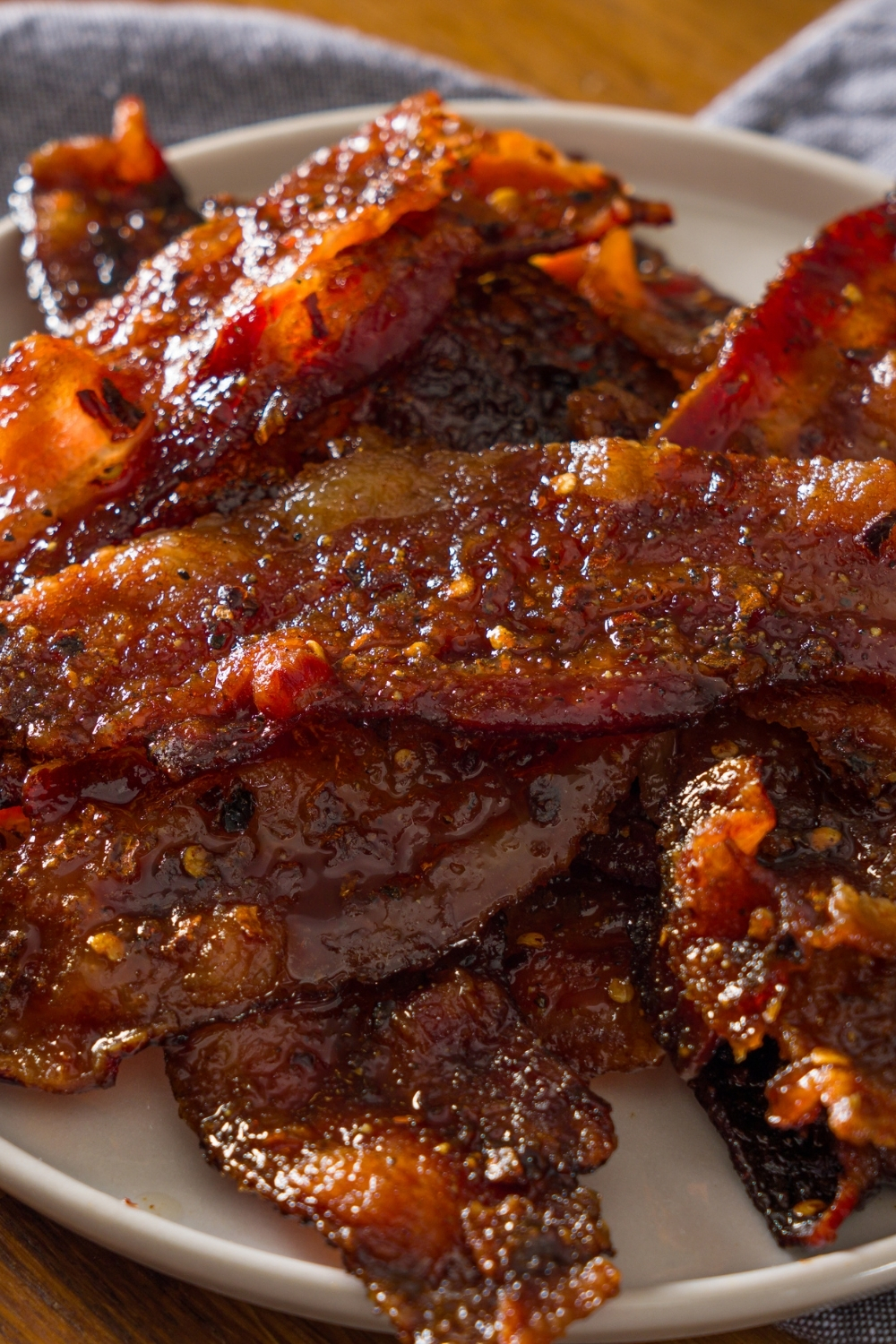 A white plate with brown sugar bacon on a wooden counter with a blue cloth napkin.