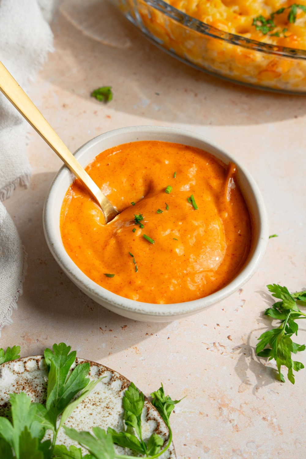 A small white bowl with campfire sauce garnished with fresh parsley. There is a spoon in the bowl. The bowl is on a tan counter with a small plate of garnishes.