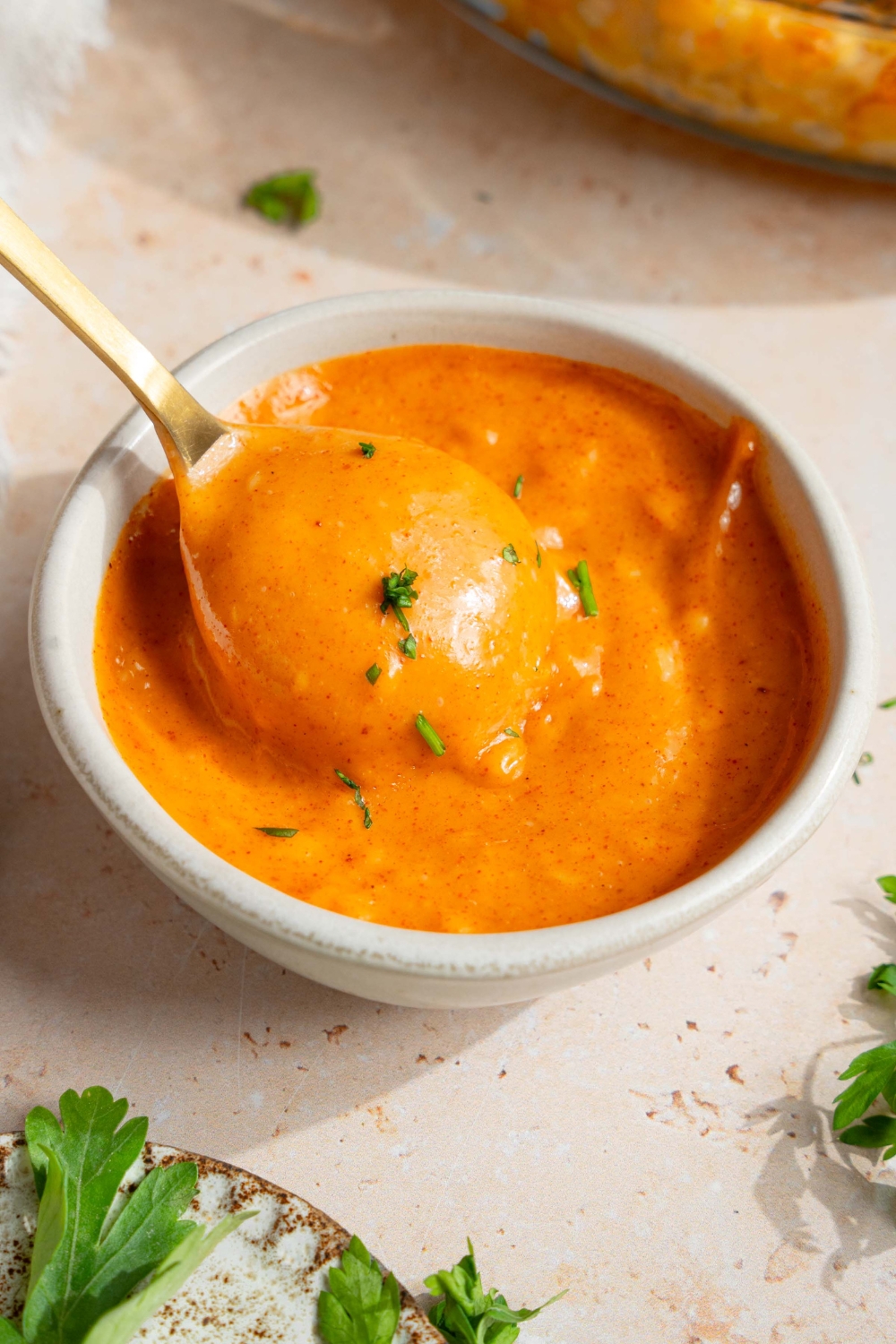 A small white bowl with campfire sauce garnished with fresh parsley. There is a spoon in the bowl. The bowl is on a tan counter with a small plate of garnishes.