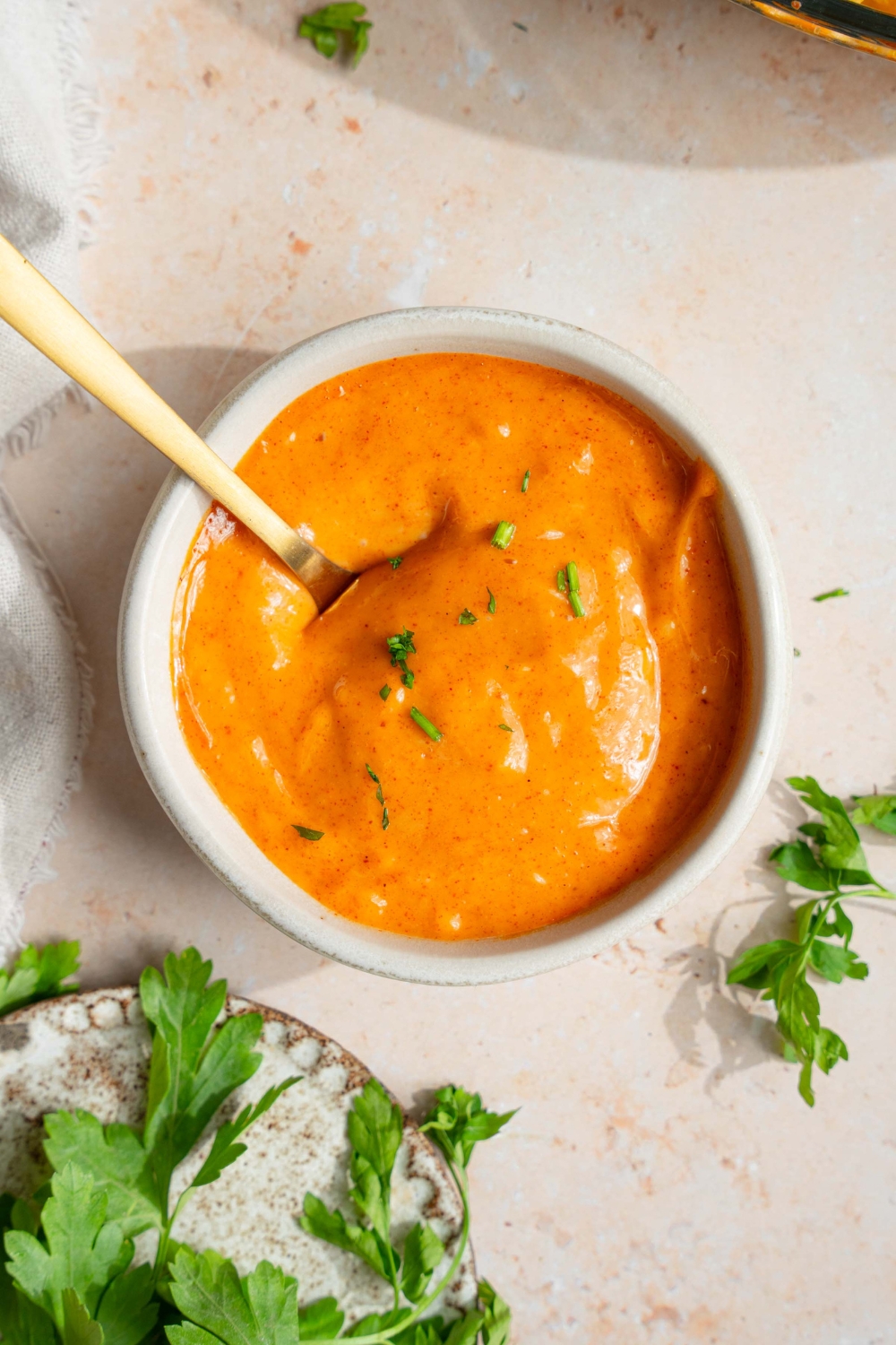 A small white bowl with campfire sauce garnished with fresh parsley. There is a spoon in the bowl. The bowl is on a tan counter with a small plate of garnishes.